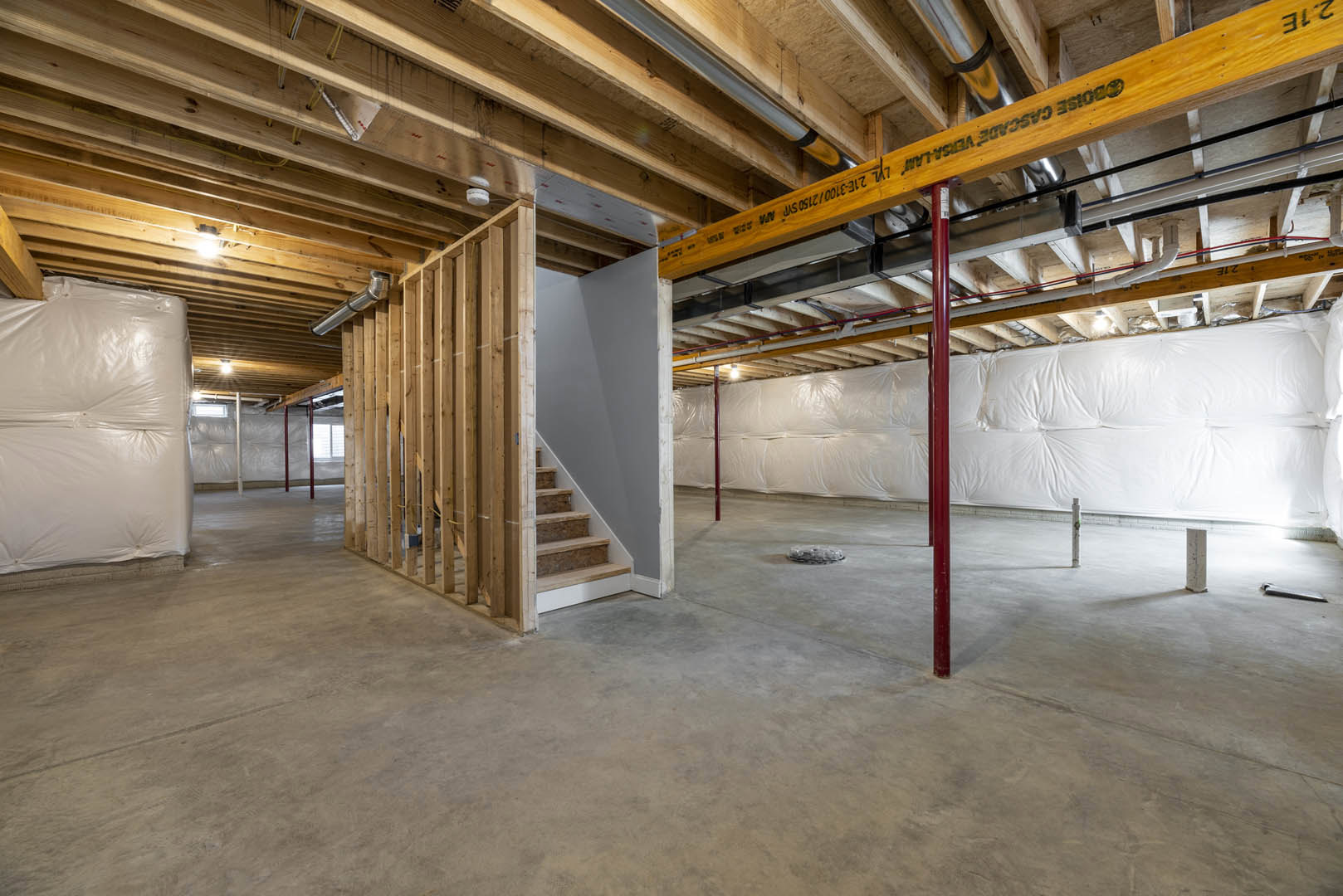 Open room with concrete floor, exposed wood beam and metal pipes overhead, white leather couch near staircase, large white bag mounted on wall, unfinished basement details visible.