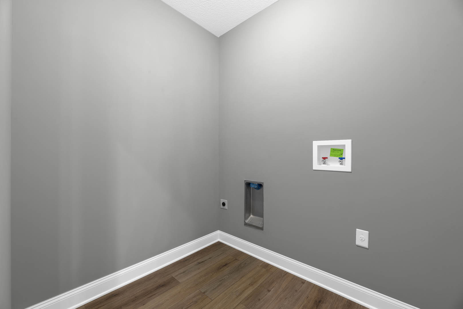 Room with light wood laminate flooring, white plaster walls, white baseboards, electrical outlet, and a small white box mounted on the wall