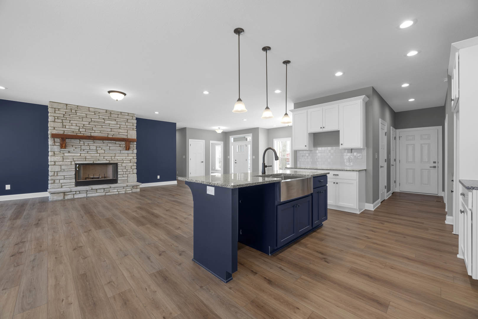 Open-concept kitchen featuring a large island with a stainless steel sink and black faucet, light cabinetry, wood flooring, a fireplace with a wood shelf and built-in bench, and a