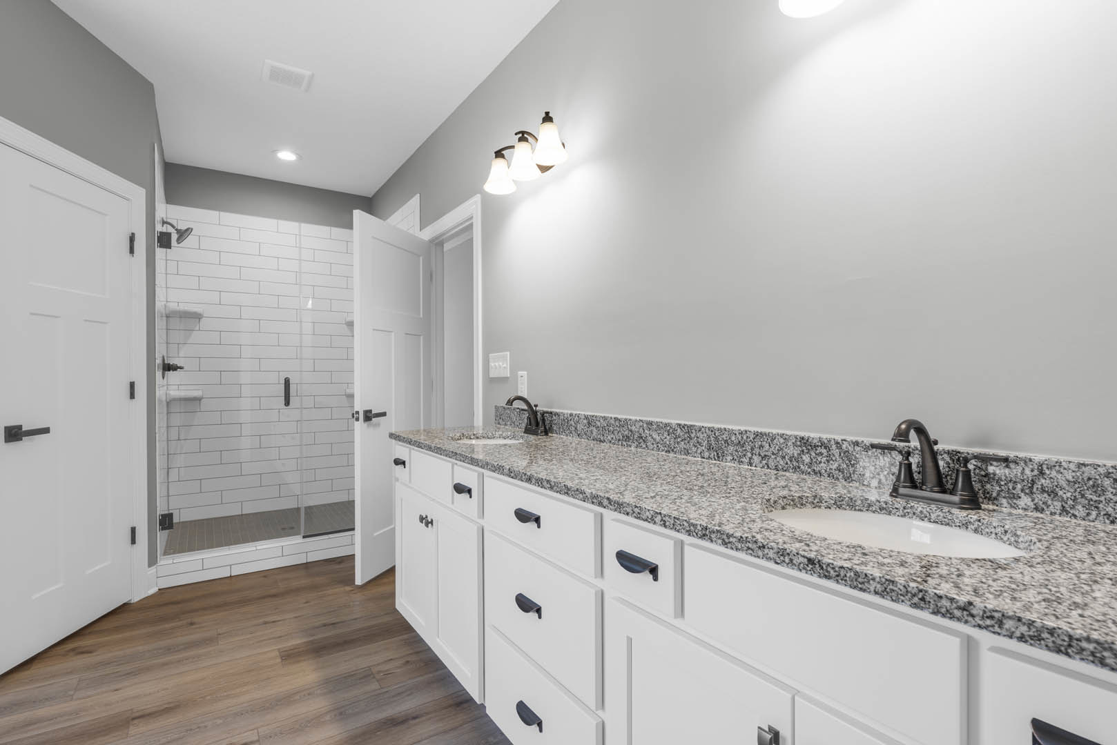 Bathroom featuring marble countertop with undermount sink, glass-enclosed shower, wood flooring, white walls, three-light fixture, white door with black handle, and modern