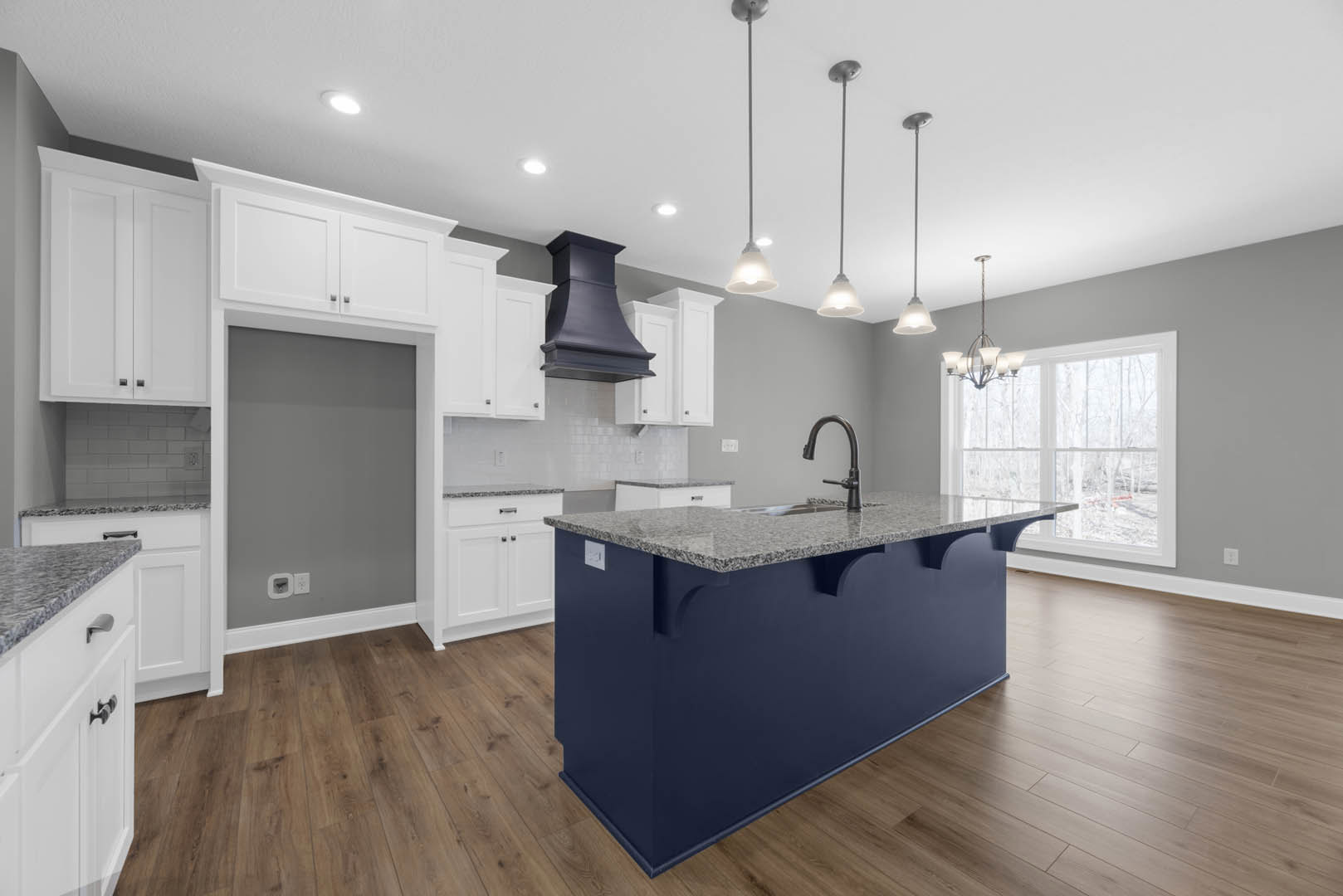 Kitchen with white shaker cabinets, granite island countertop, stainless steel sink and faucet, large window above counter, light wood flooring, modern appliances, and built-in