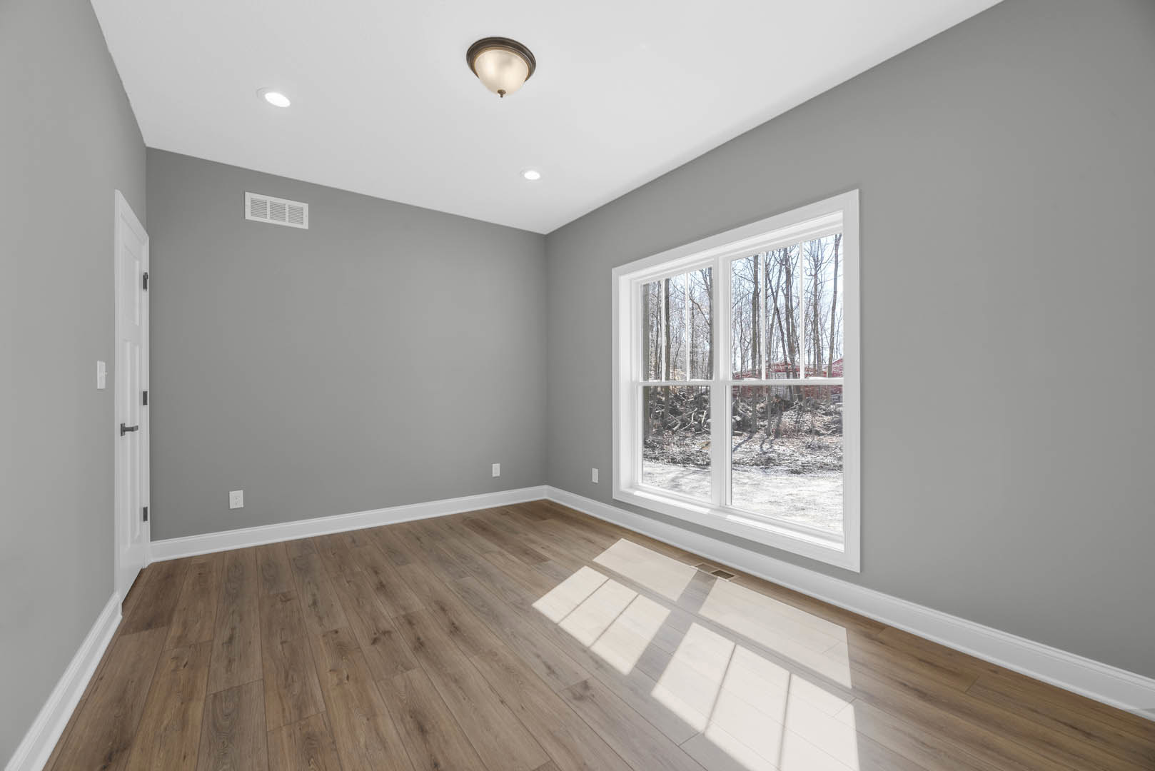 Sunlit room featuring wide plank wood flooring, large window with tree views, white plaster walls, ceiling-mounted light fixture, and decorative molding.