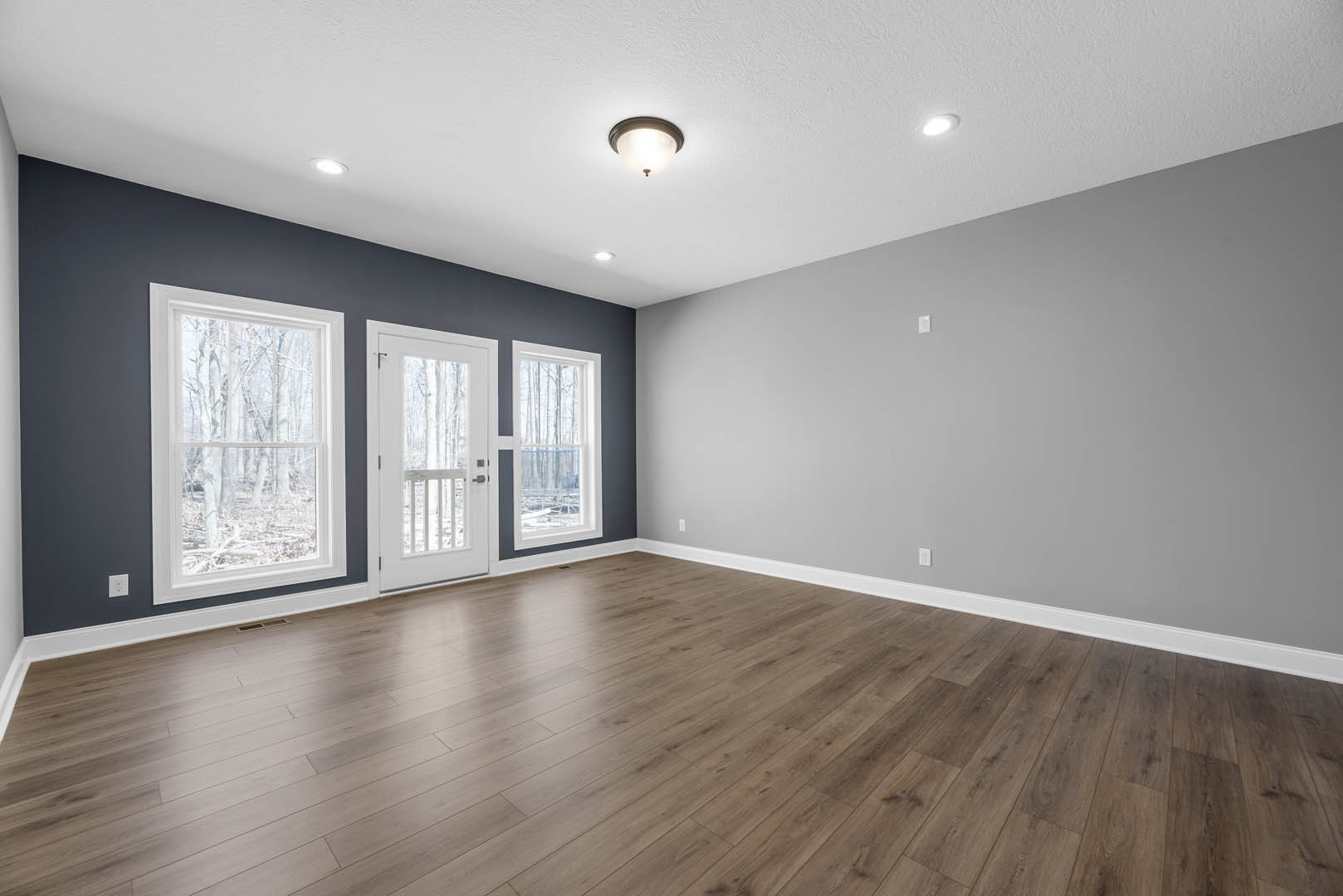 Wood floor with natural finish, white plaster walls, large window framed in white showing trees outside, white door with glass panel, ceiling-mounted light fixture