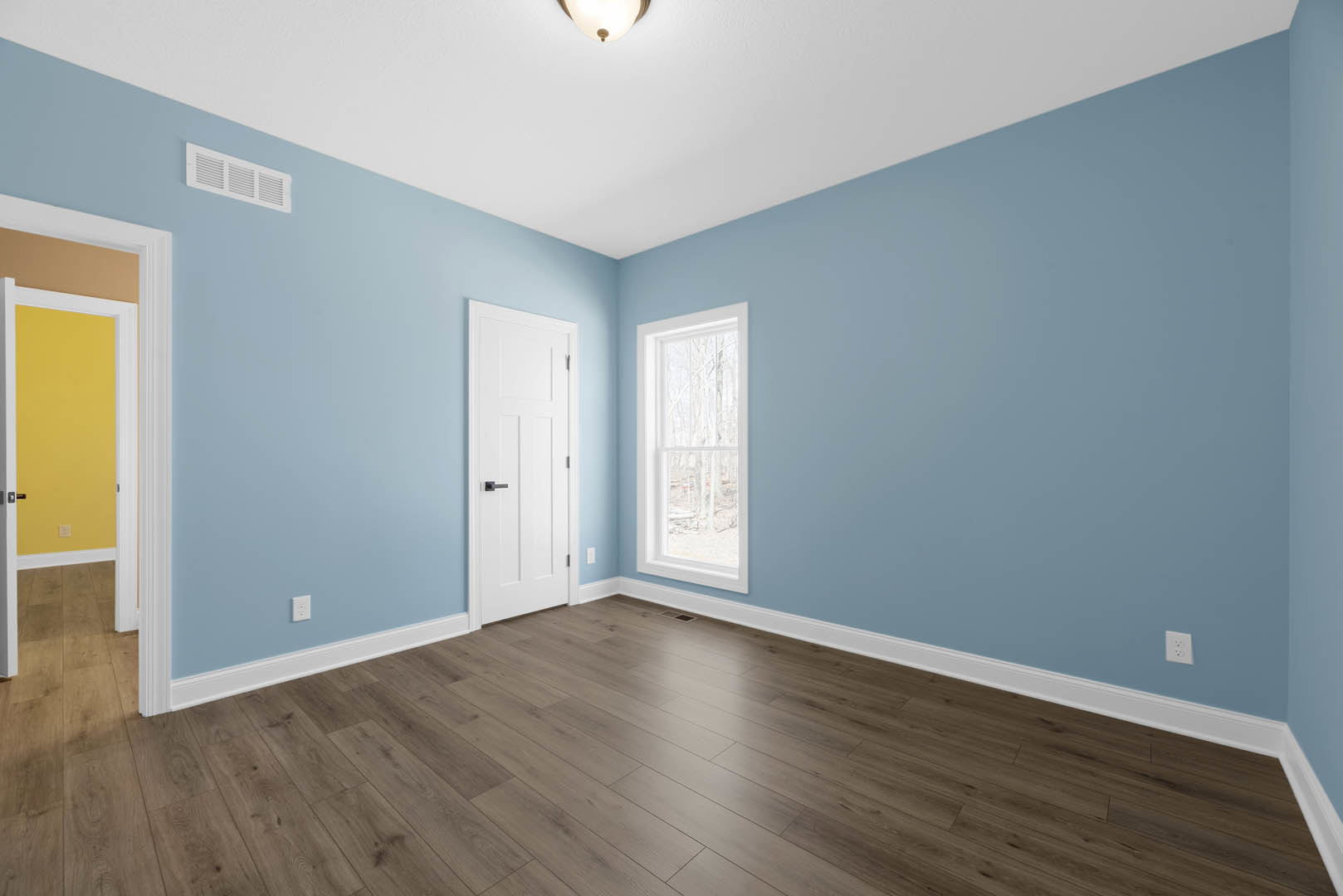 White walls and wood flooring in a room with a white door featuring a black handle, a window overlooking trees, a yellow rectangular object with a white border, and a wall vent.