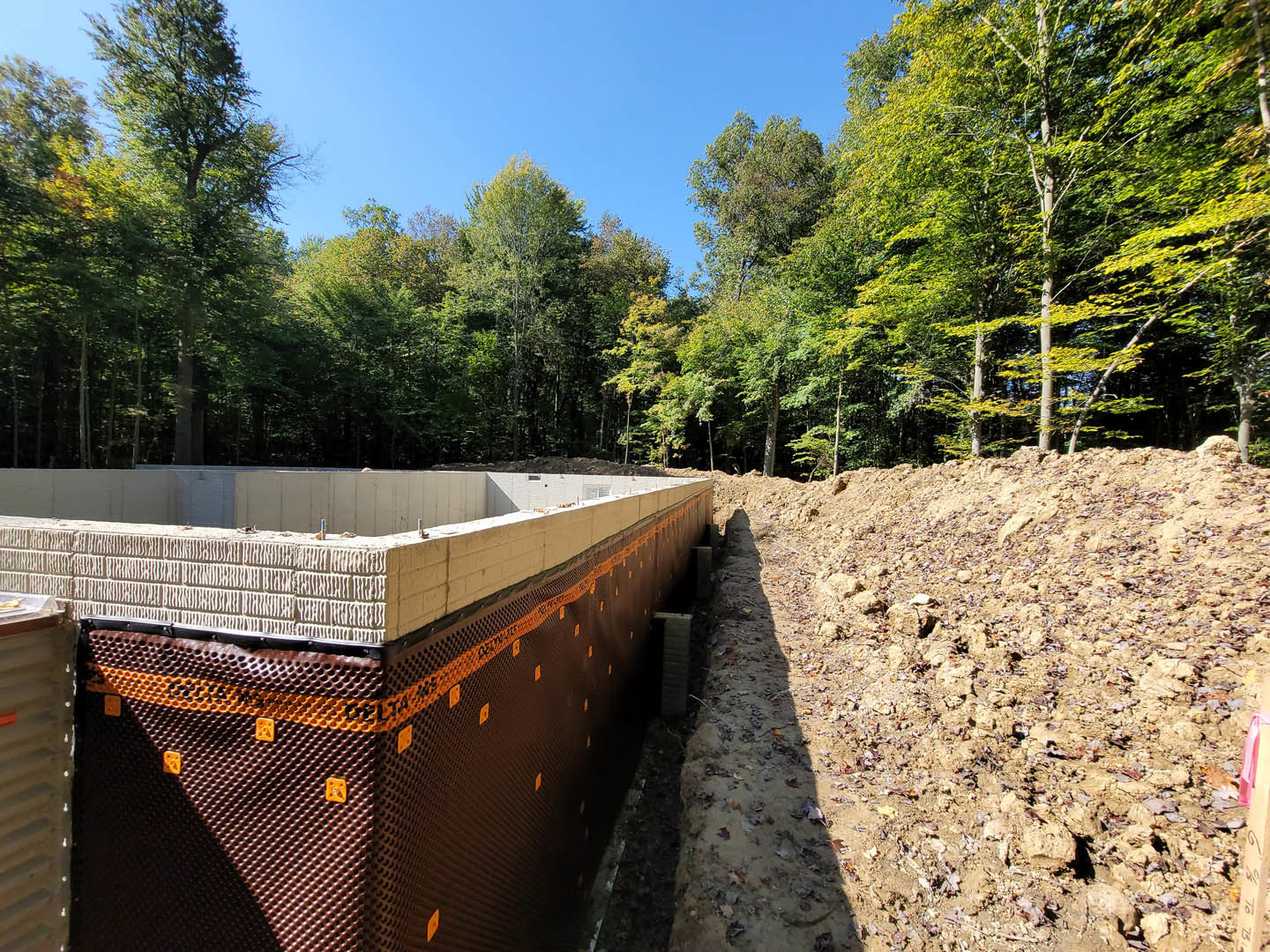 Concrete foundation with brick wall and foam insulation, dirt hill in foreground, forested area and trees in background, partial window visible