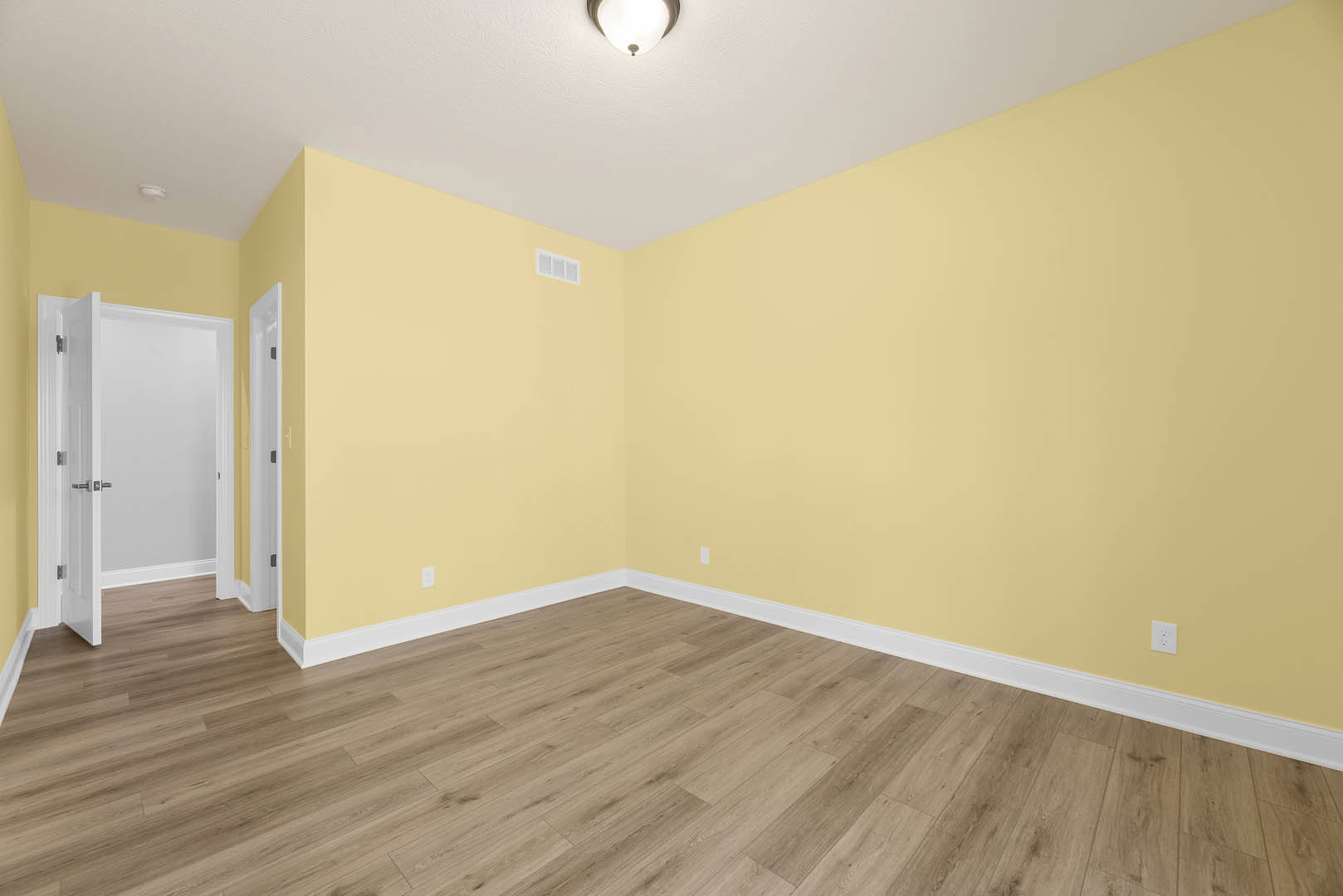 Yellow walls, wood flooring with white baseboards, white door with silver handles, close-up of ceiling light, white wall vent