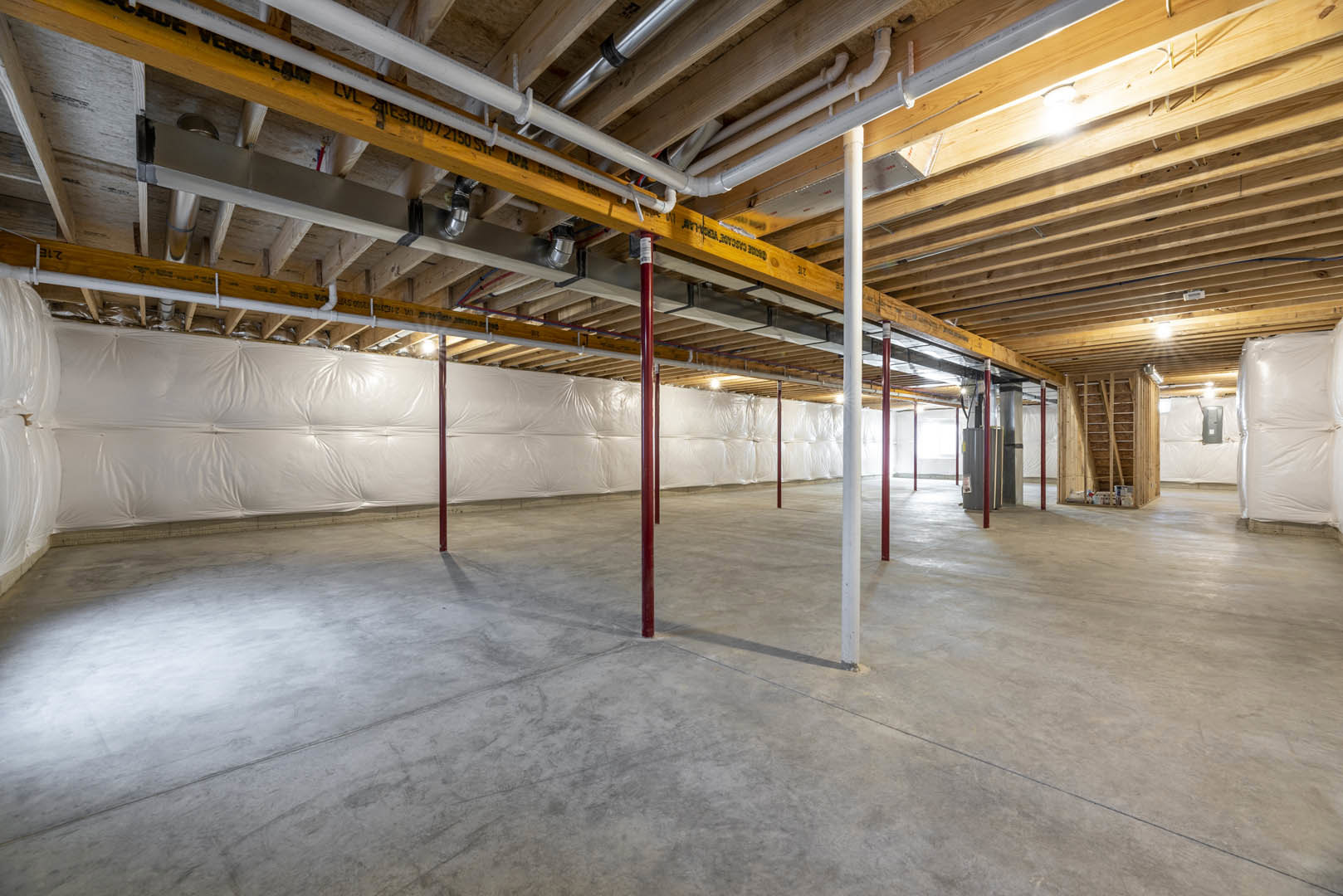 White-walled room with exposed wooden ceiling beams, concrete floor, red support poles, white leather couch, and visible ceiling pipes.