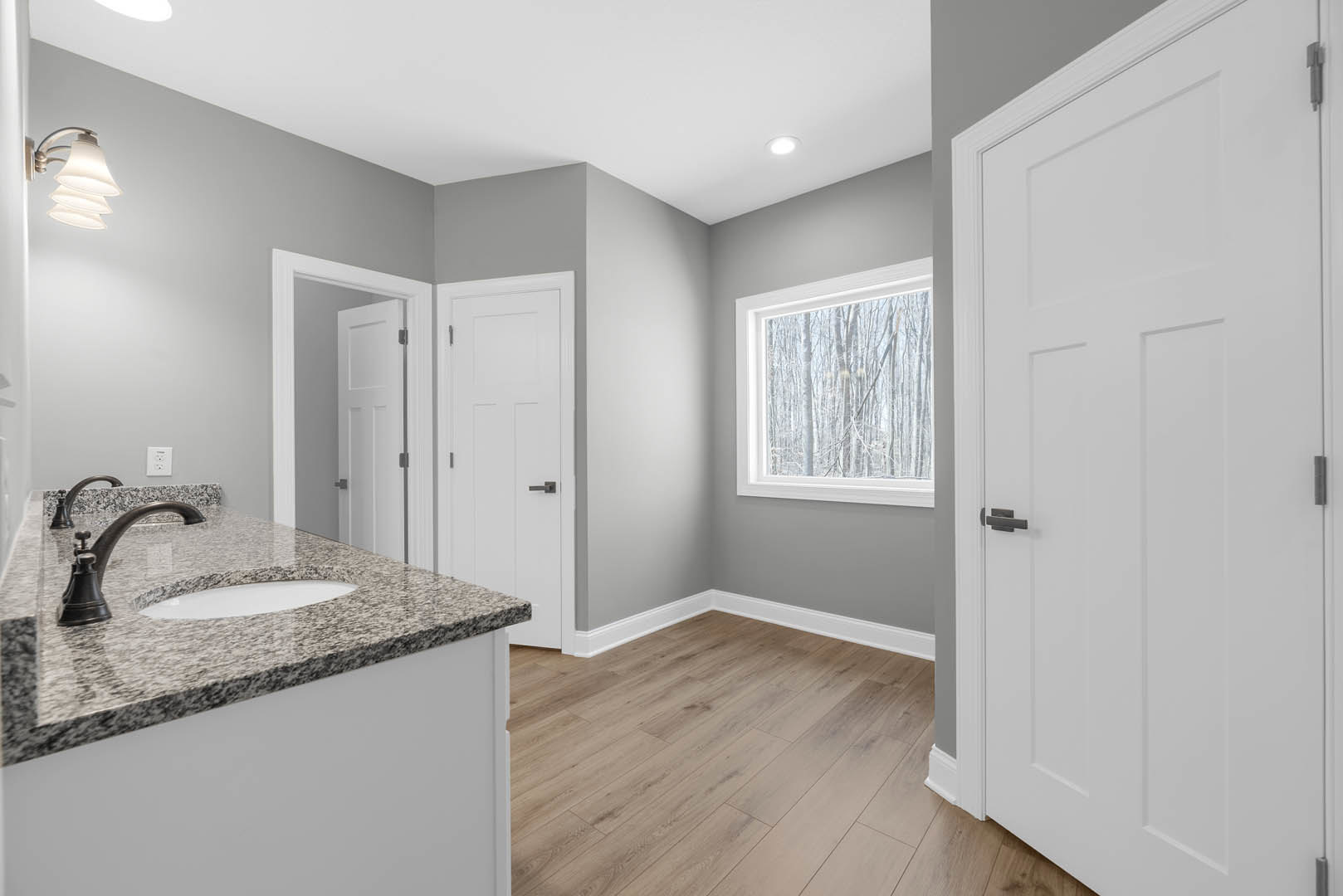 Bathroom featuring marble countertop, wood flooring, white sink with black and white counter, black-handled white door, window overlooking trees, and white-shaded light fixture