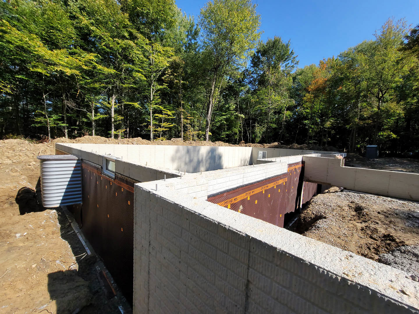 Partially built exterior wall with exposed concrete foundation, white plumbing pipe, blue trash can, and surrounding trees in the background