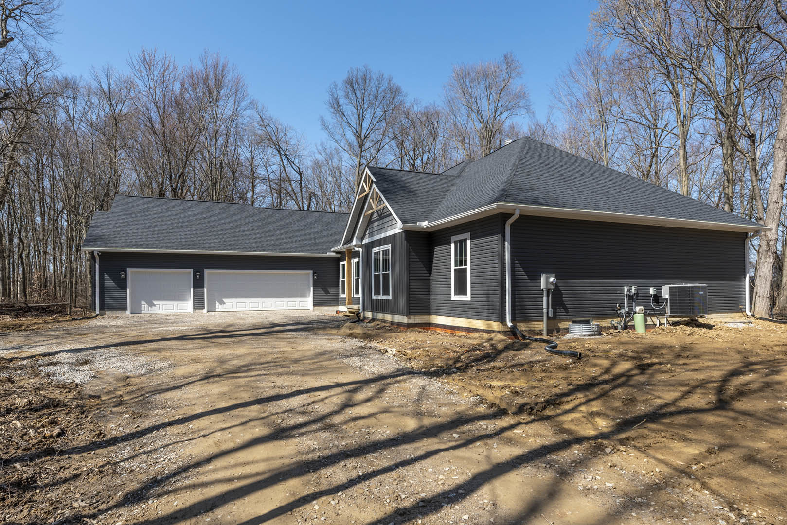 Black siding house with white garage door, black roof, paved driveway, and mature trees in the background