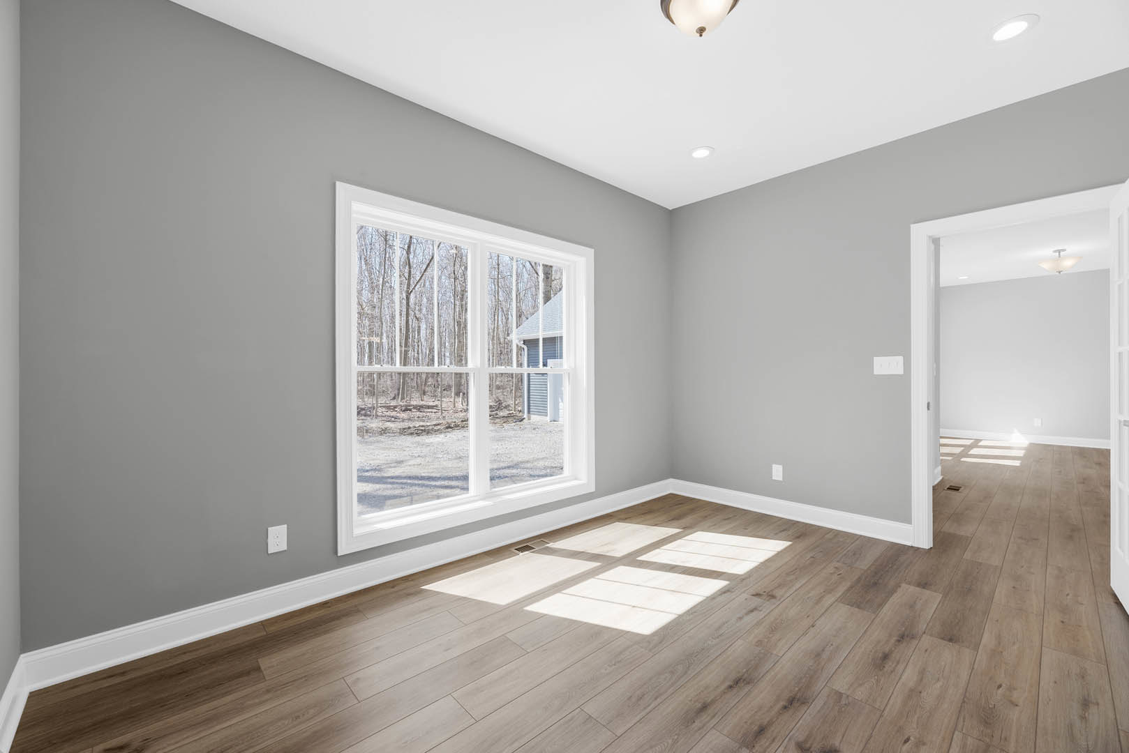 Sunlit room featuring wide wood plank flooring, large window with views of neighboring house and trees, smooth white walls, and recessed ceiling light.