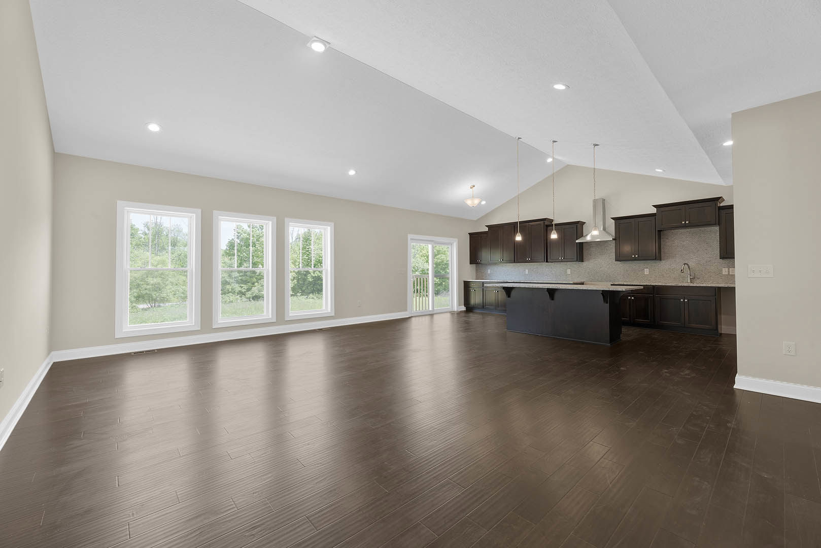 Spacious open-plan room featuring a central kitchen island, wide plank wood flooring, white ceiling with recessed lights, and a row of large windows framed in white overlooking