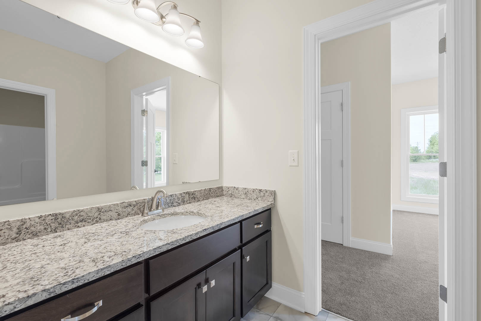 Bathroom with marble countertop, undermount sink, white cabinetry, large mirror, white walls, and door; window reveals trees outside.