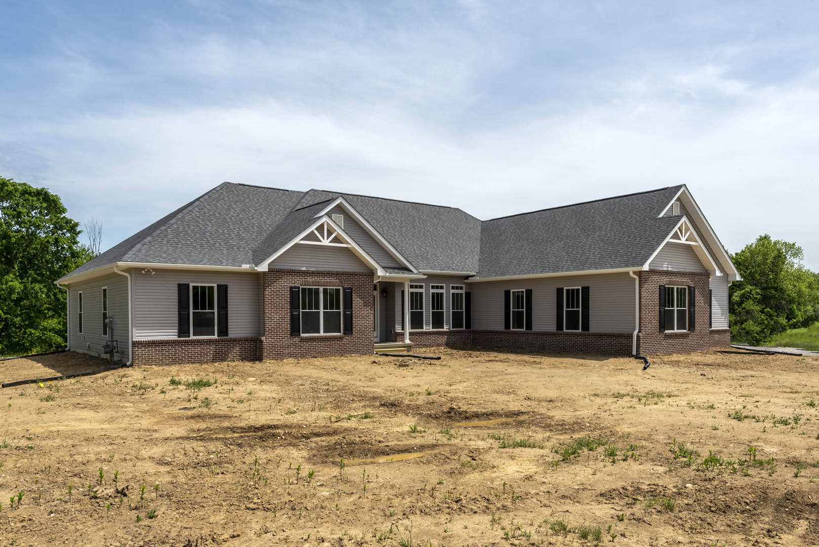 Brick house with black shuttered windows, glass-paneled door, and brick roof, set beside a dirt field with a hole, surrounded by trees under a cloudy sky