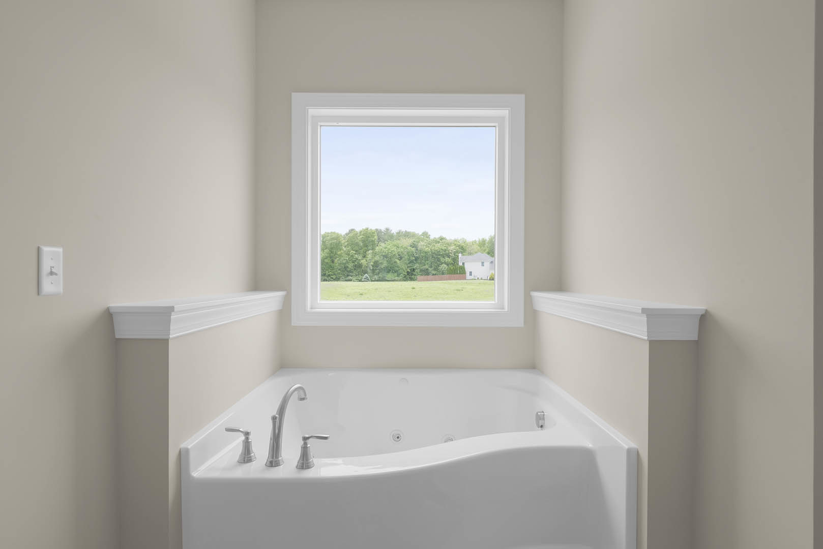 Freestanding white bathtub beneath a large window overlooking a field and trees, surrounded by white tile, wall molding, and a built-in shelf