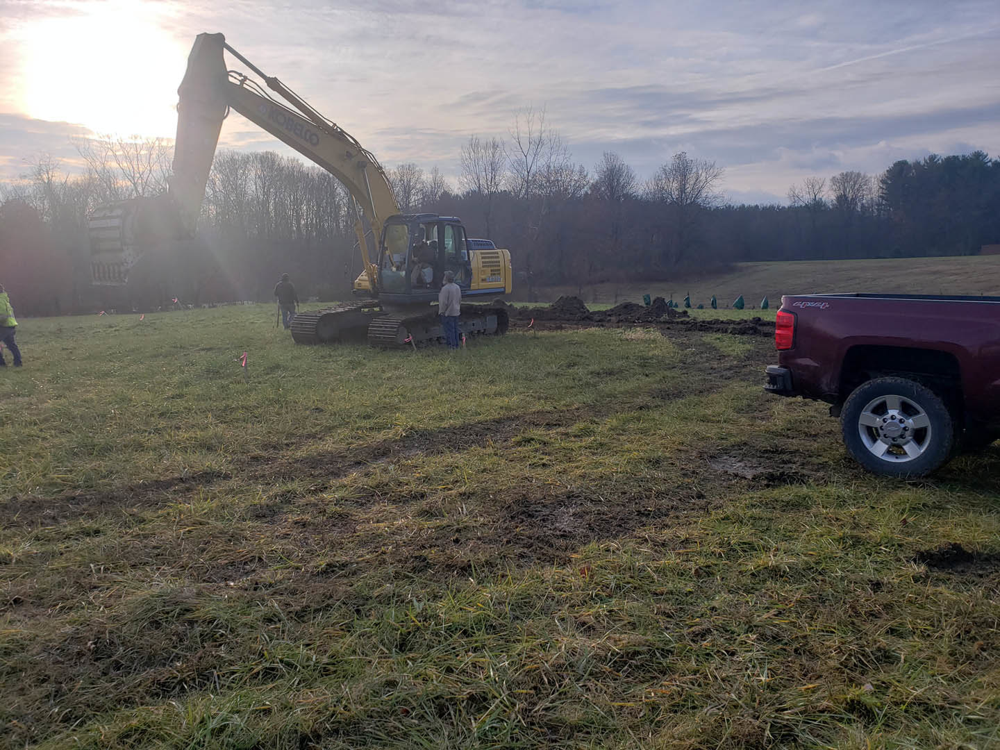 Bulldozer and construction crew on grassy field with cloudy sky, trees in background, close-up of tire and truck visible