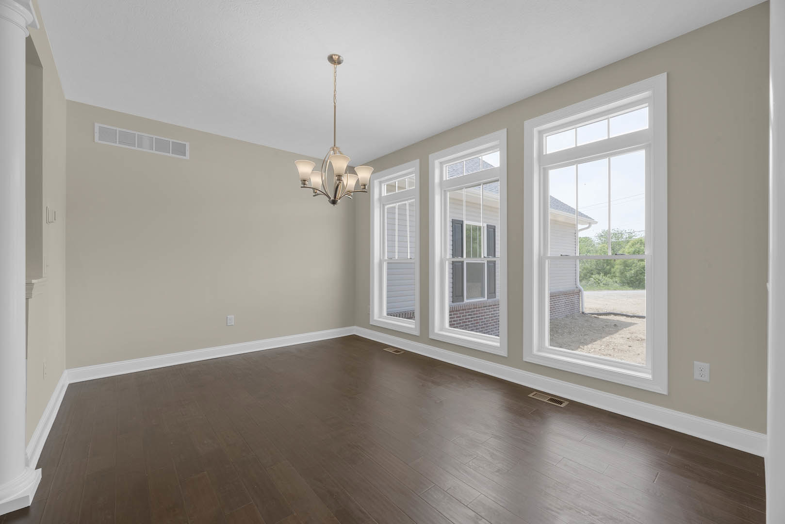 Chandelier with white shades hanging above hardwood flooring, large windows with outdoor view, white plaster walls and crown molding