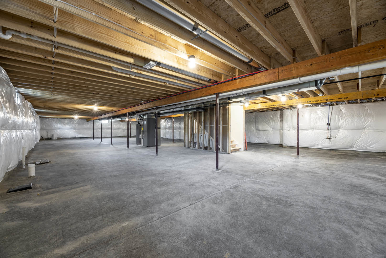 Concrete floor with visible crack, exposed wood beams, white wall partially covered in plastic sheeting, stack of white plastic bags, ceiling illuminated by overhead light.