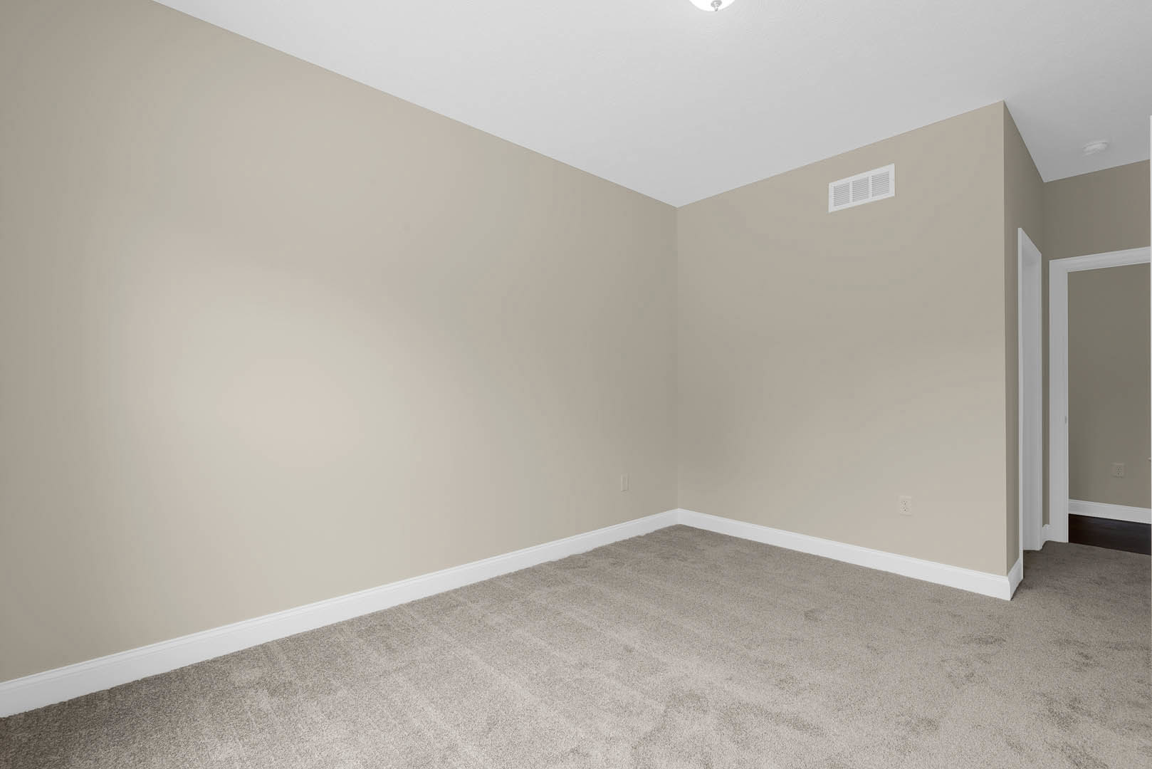 Beige walls and carpeted floor in a residential room, white baseboards, wall vent, closed door visible