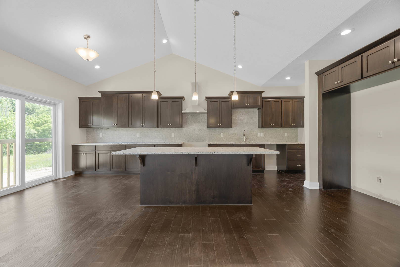 Spacious kitchen featuring a large central island with stone countertop, dark wood flooring, sleek cabinetry, pendant light fixture, and a window overlooking leafy trees.