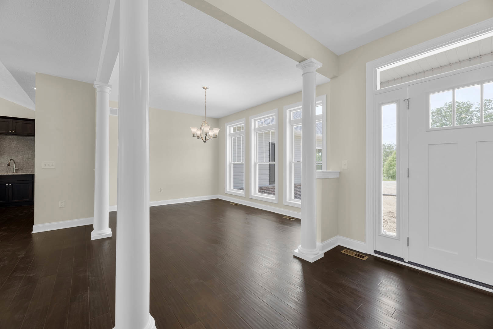 Room with white pillars, dark wood flooring, hanging light fixture, white door with glass panes, and window with molding