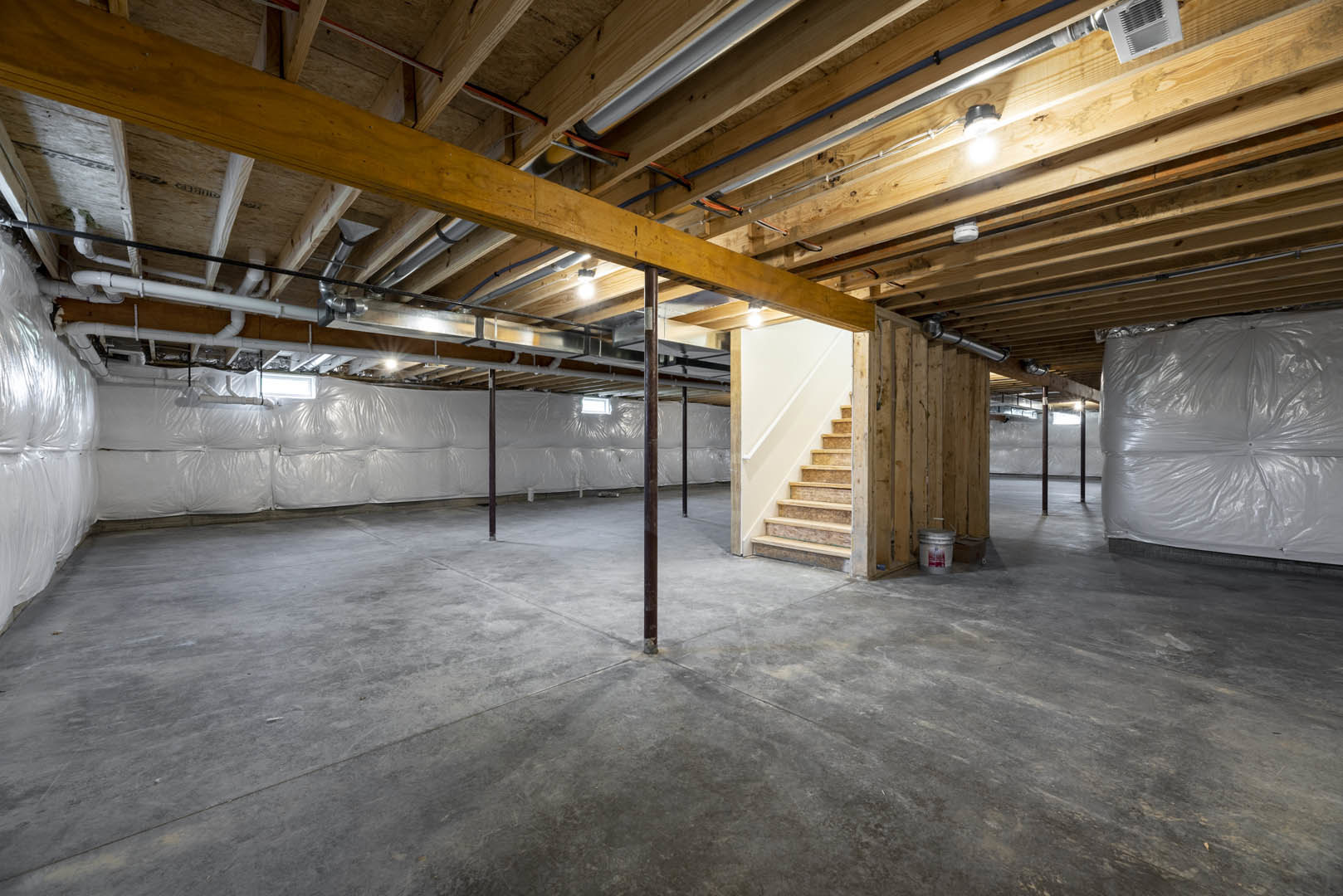 Basement with exposed wooden beam ceiling, staircase, concrete floor with metal pole, white plastic bag, red-labeled paint bucket, visible ceiling pipes, and close-up vent.
