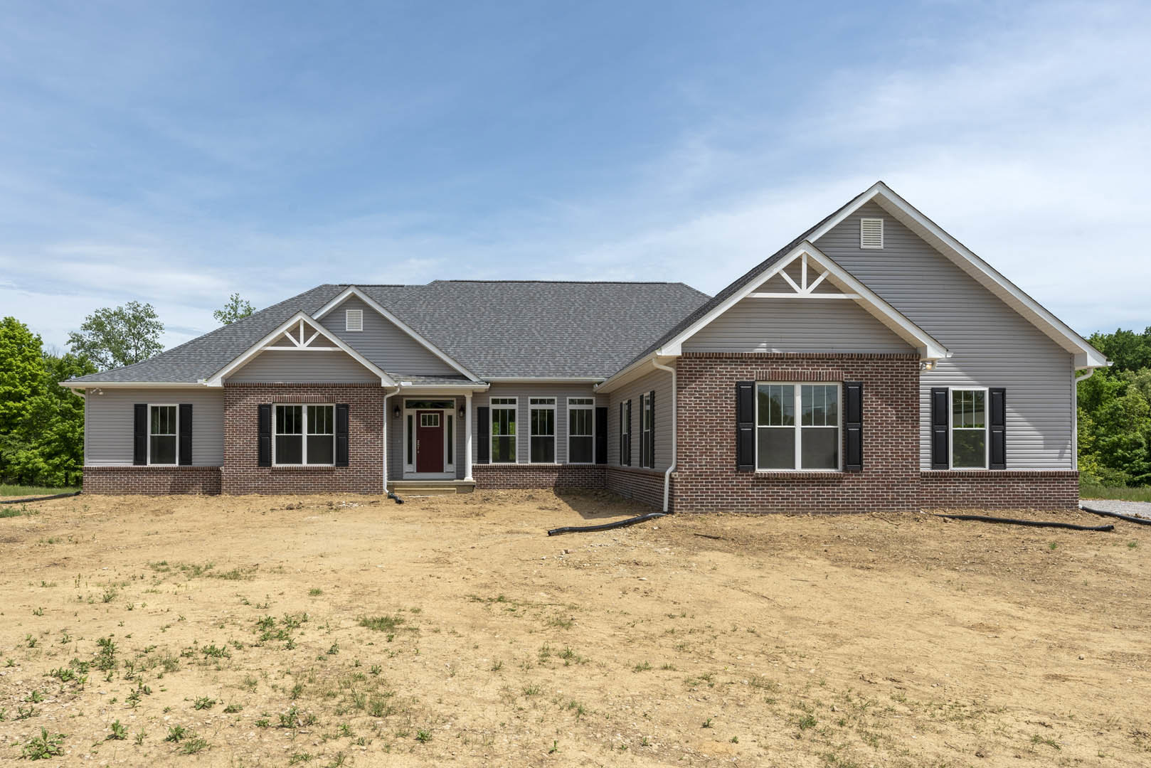 Two-story house with white siding, red front door, double windowed doors, black pipe lying on bare dirt yard, cloudy sky overhead, trees in background.