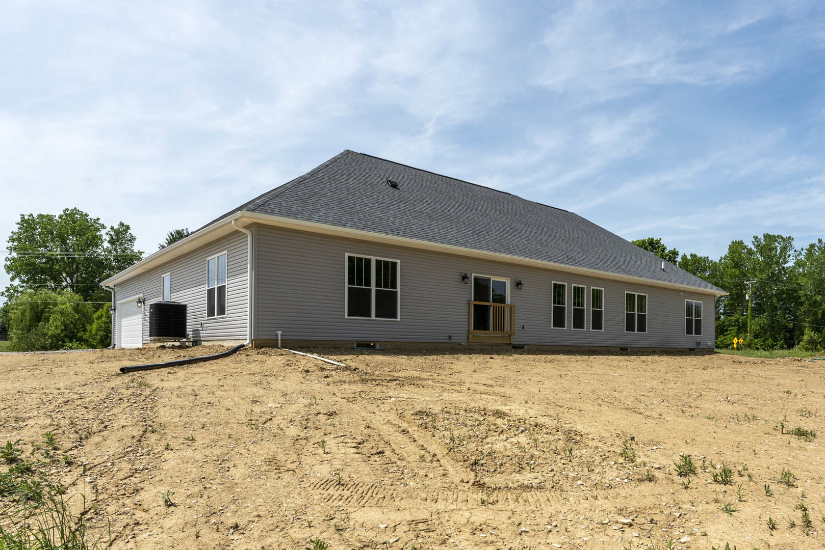Single-story house with white-trimmed windows, wooden railing, and black square object on metal stand beside dirt field marked with tire tracks under partly cloudy sky