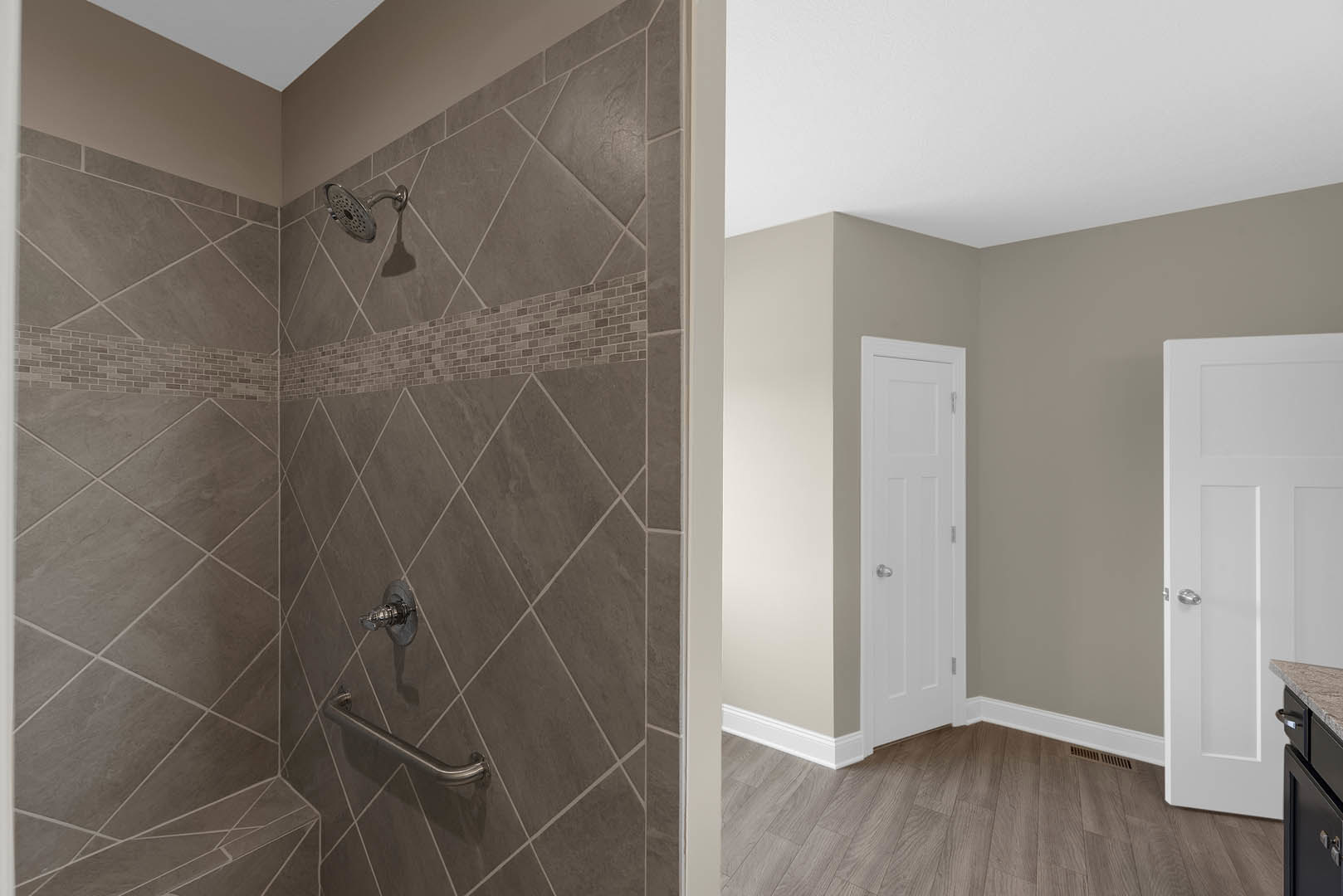 Modern bathroom with light tile walls, glass shower enclosure featuring a hand shower, white paneled door with silver knob, black vanity drawer, and chrome handrail.