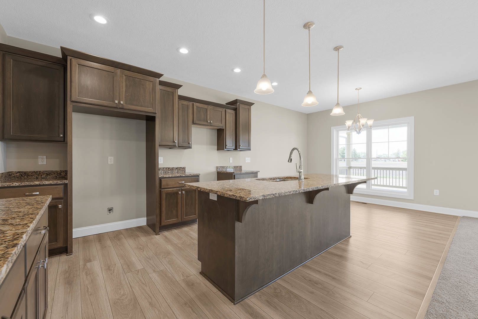 Spacious kitchen featuring a large central island with stone countertop and built-in sink, brown cabinetry, white walls with electrical socket, oversized window, modern light