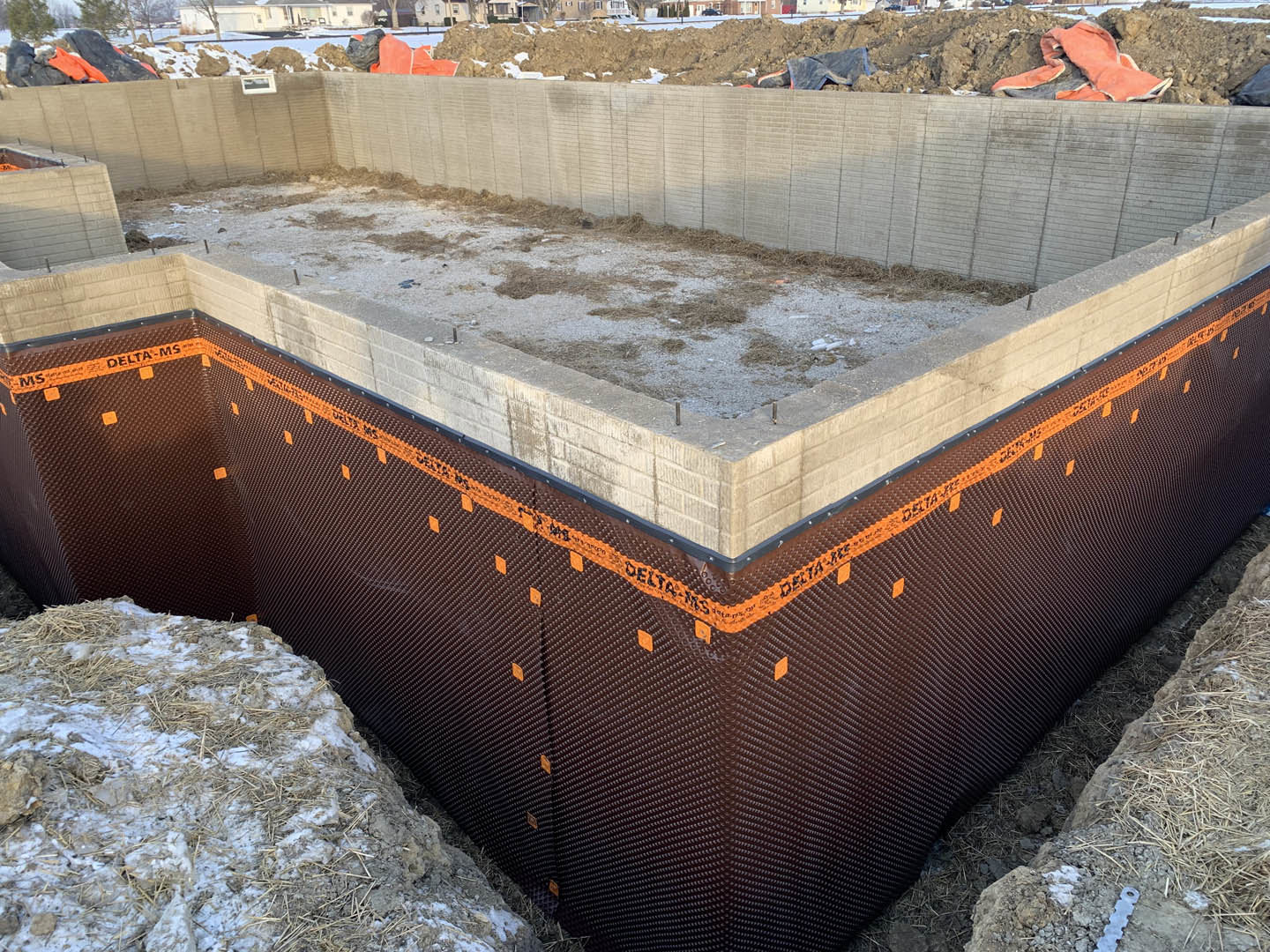 Concrete foundation slab with exposed rebar, surrounded by dirt, straw, and patches of snow on a residential construction site