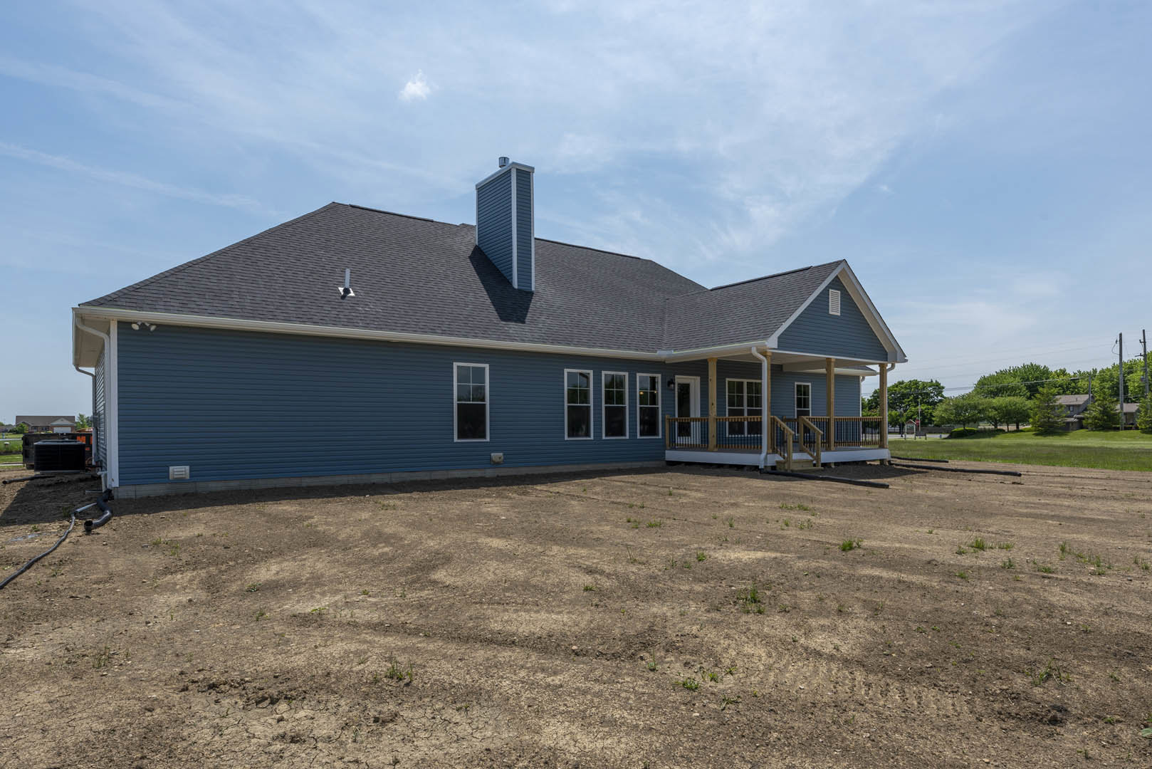 White-sided house with prominent chimney, large window, and expansive grassy yard bordered by dirt patches and mature trees under a partly cloudy sky