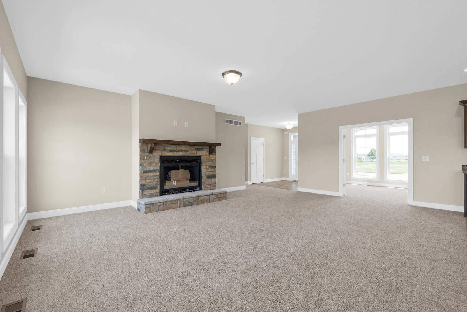 Living room with beige carpet, white walls, brick fireplace containing a cardboard box, ceiling light fixture, open door, and window letting in natural light