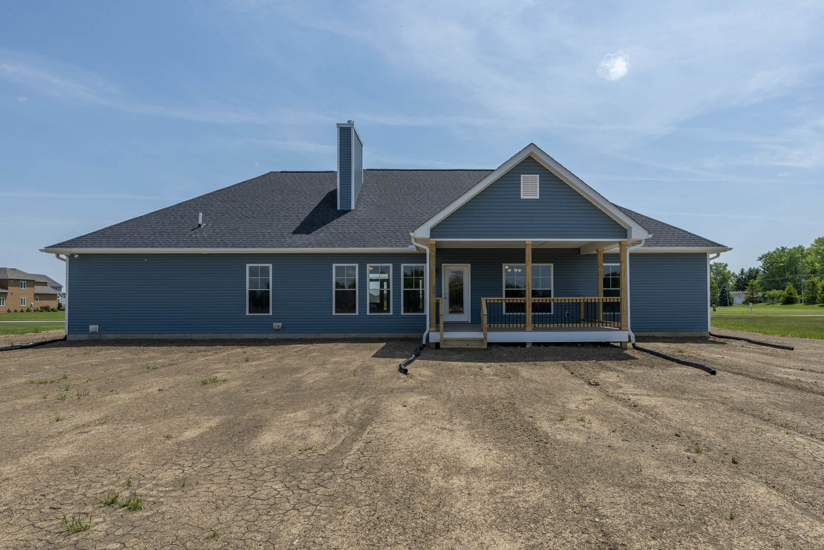 Blue cottage-style home with white door, covered porch, white-framed windows reflecting neighboring house, exposed drainpipe, and bare dirt yard under partly cloudy sky.