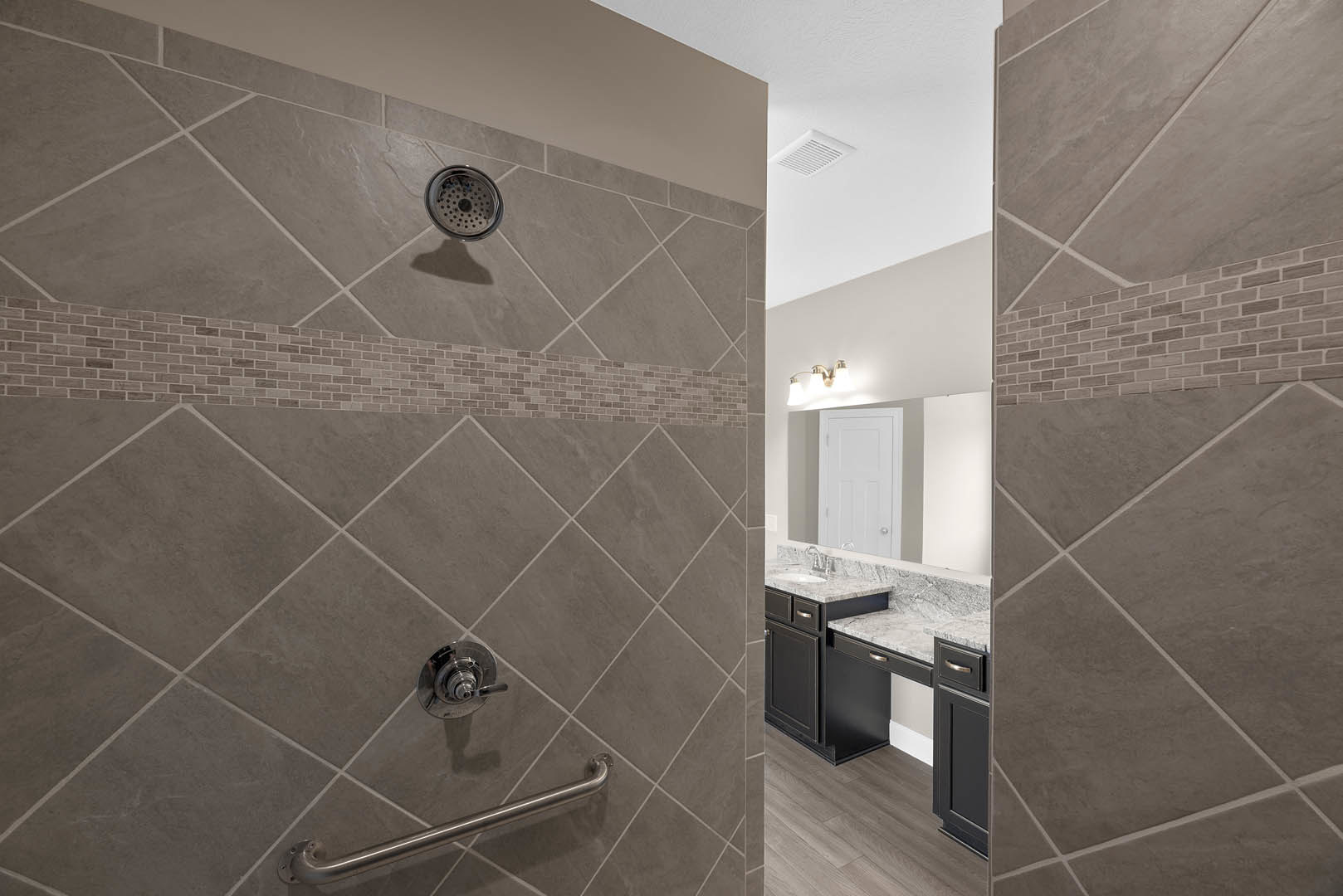 Walk-in shower with large gray tile walls, chrome shower head and hand shower, glass door, metal grab bar, and tile floor with linear drain