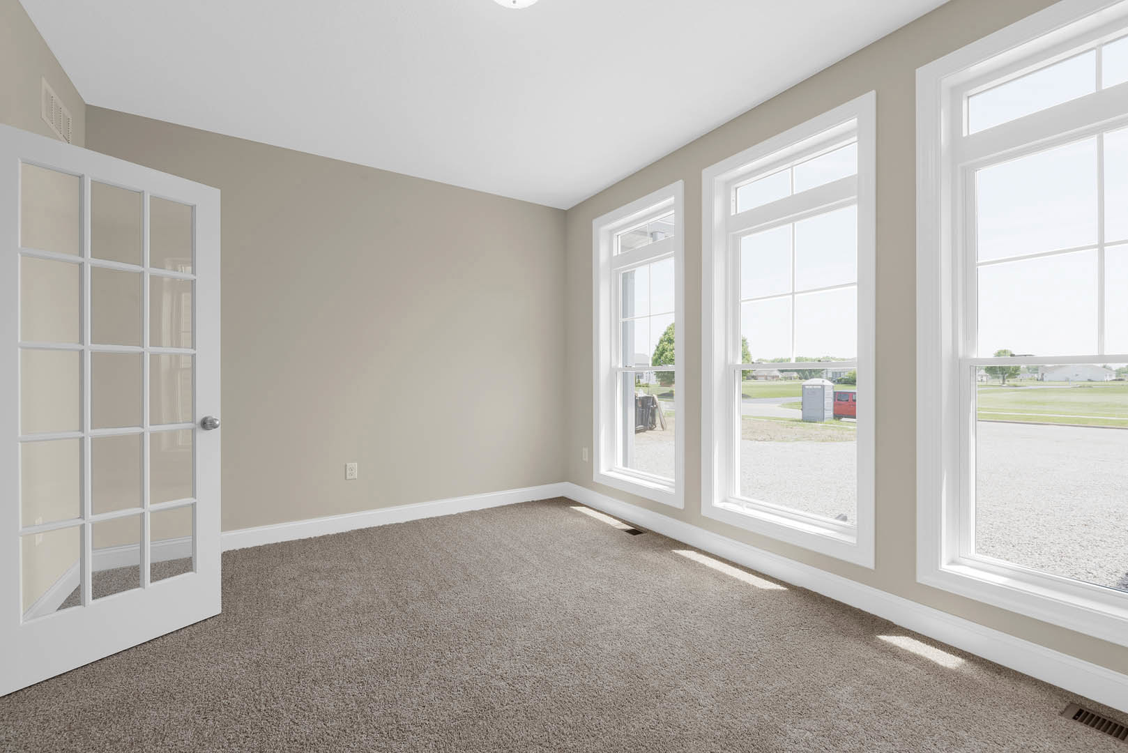 Spacious room with multiple large windows, white walls, light carpet flooring, and a single door opening to a grassy outdoor area