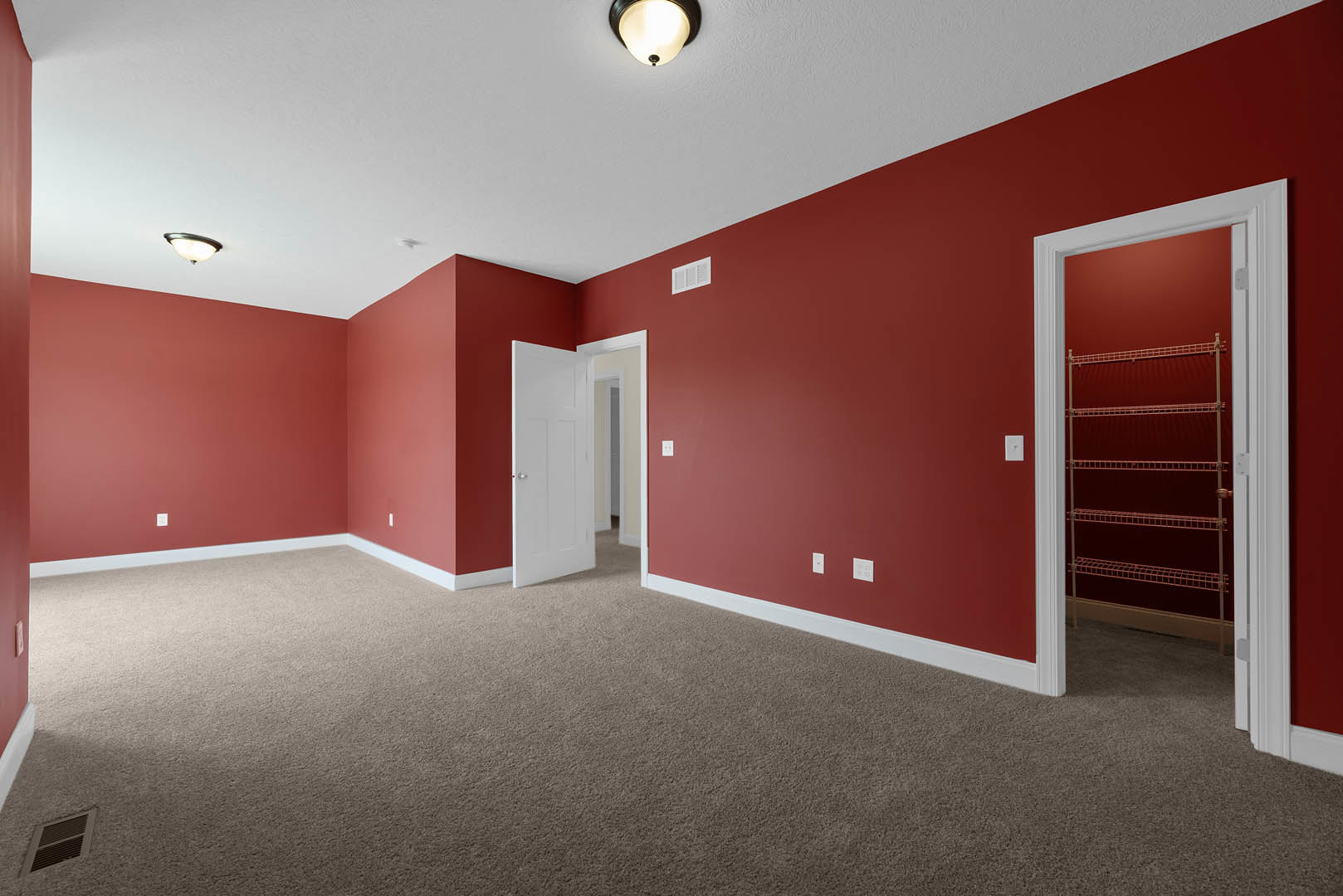 Red walls and red carpeted floor in a room with a white ceiling vent, a close-up of a ceiling light, a door with a built-in shelf, and a red metal wall shelf.