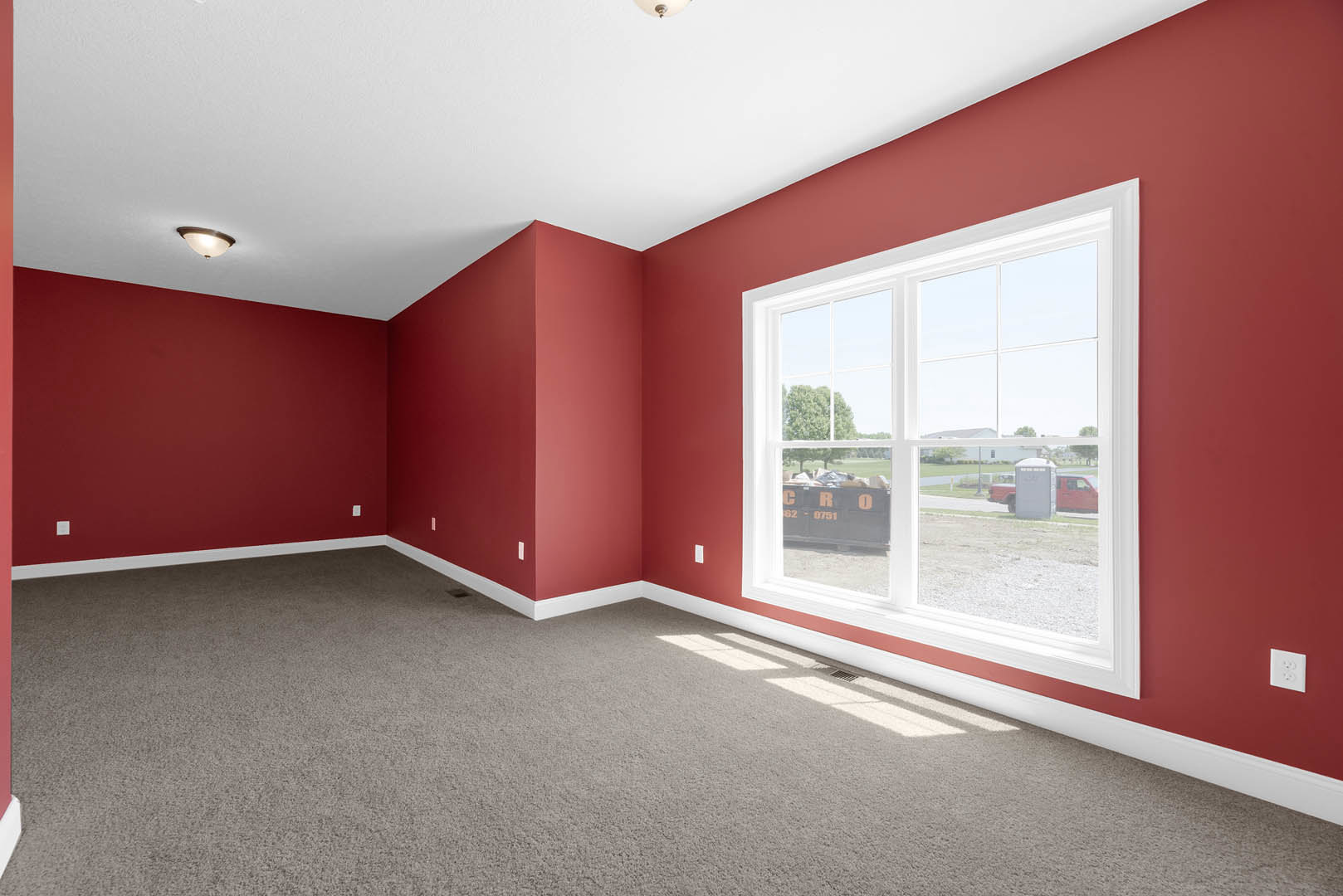 Red-walled room with red carpet, white electrical outlet, window showing passing train, ceiling light fixture illuminated