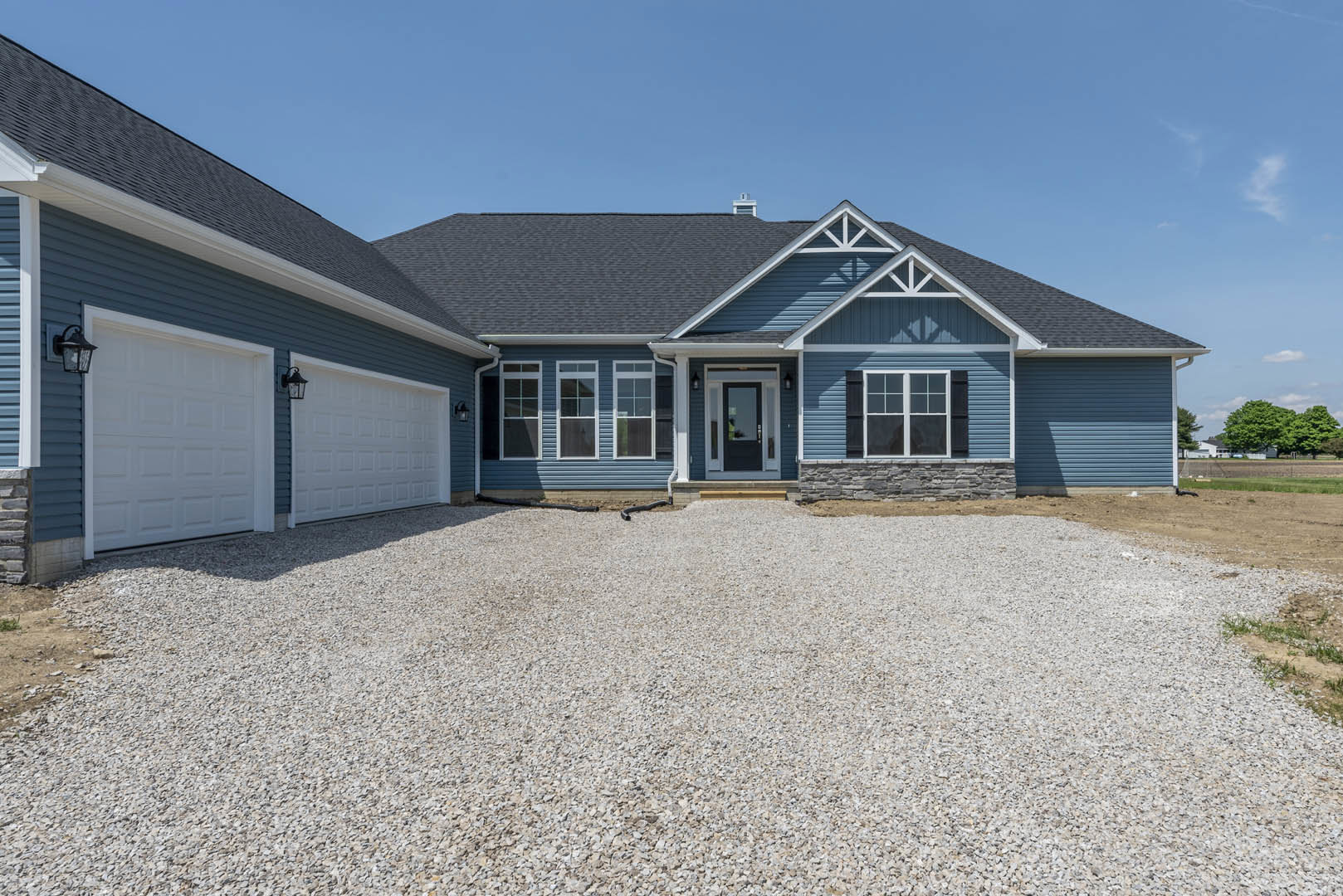 Gravel driveway leading to a white garage door with lamp post, black door with glass panes, white-framed window, and black hose on ground