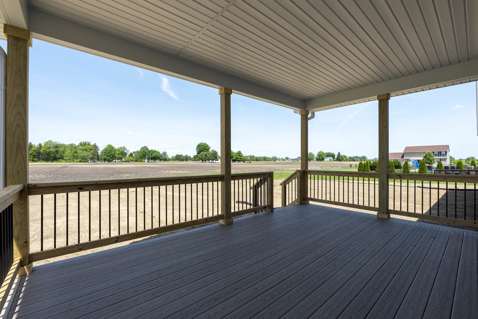 Wooden deck with railing, bordered by a fence, overlooking a field and trees; white roof supported by exposed wooden beams.