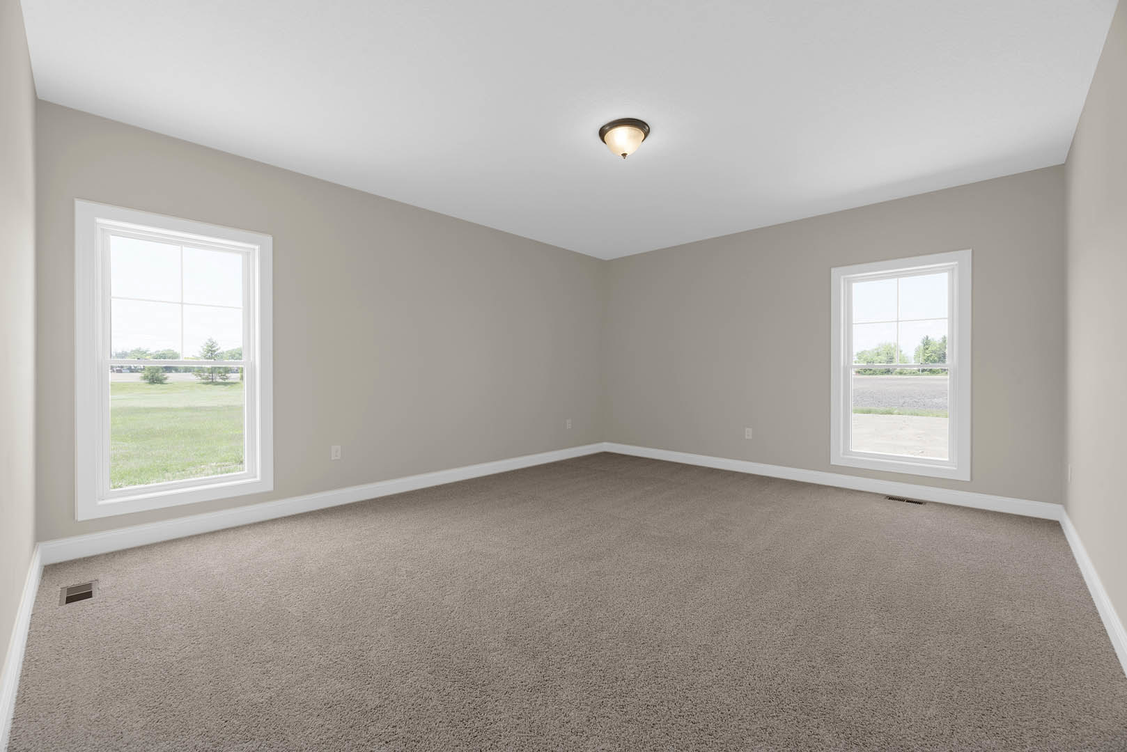 Carpeted room with white walls, large window overlooking grassy field and road, ceiling light fixture, simple baseboard molding