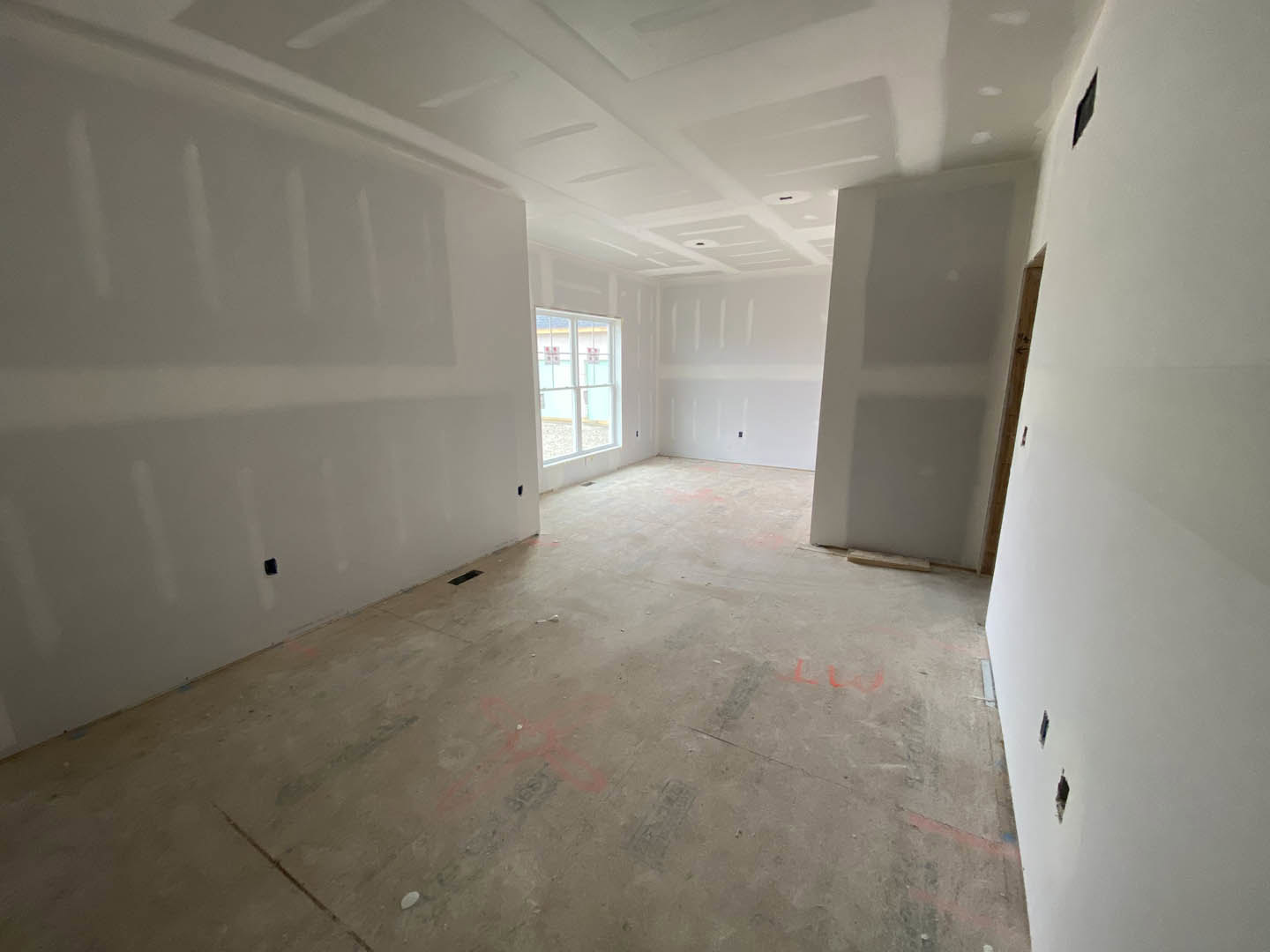 White plaster walls, large window with sunlight, concrete floor partially painted red, red and black fan on floor, black holes visible in wall.