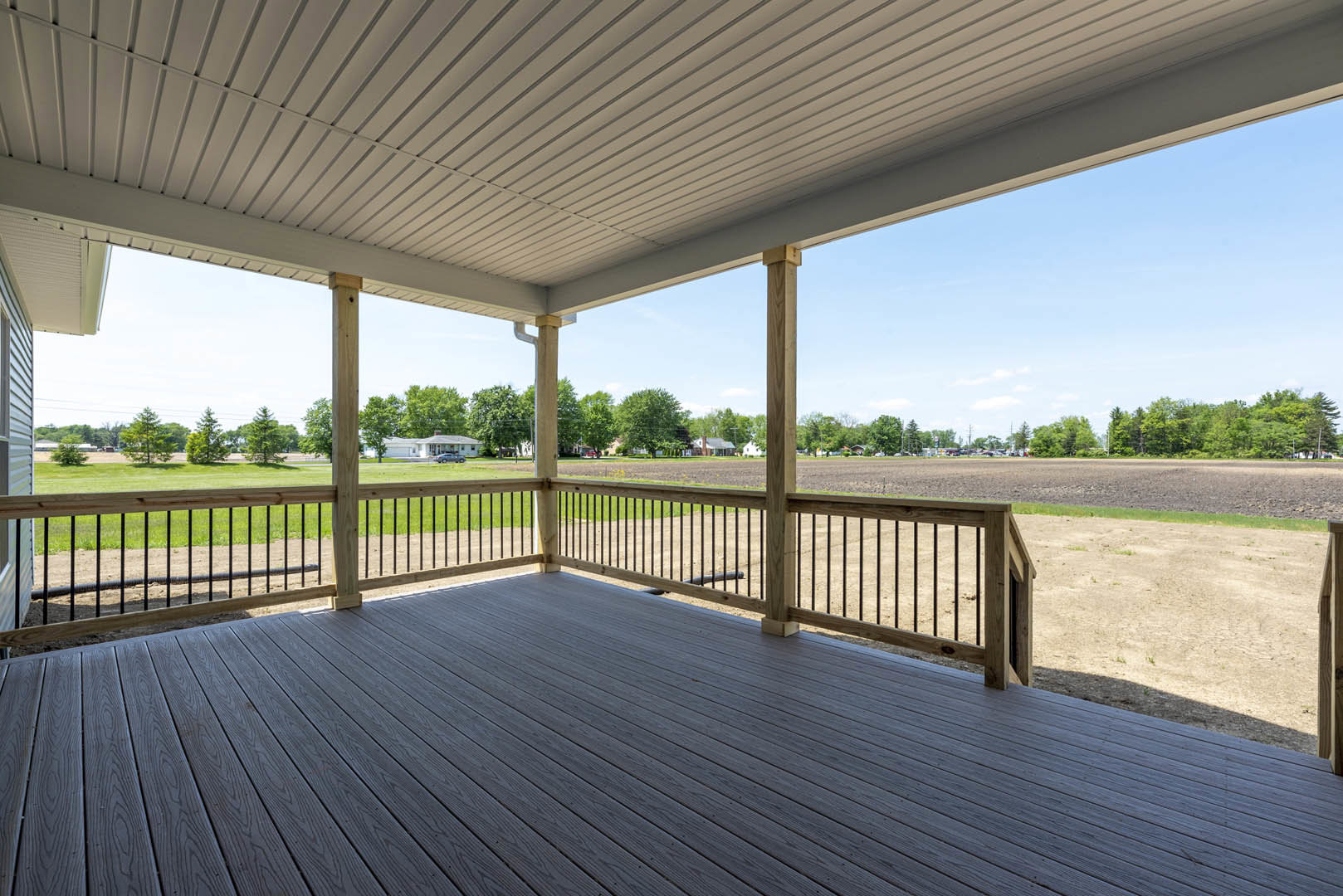 Wooden deck with railing overlooking grassy field, white roof supported by exposed wood beams, group of trees in the background