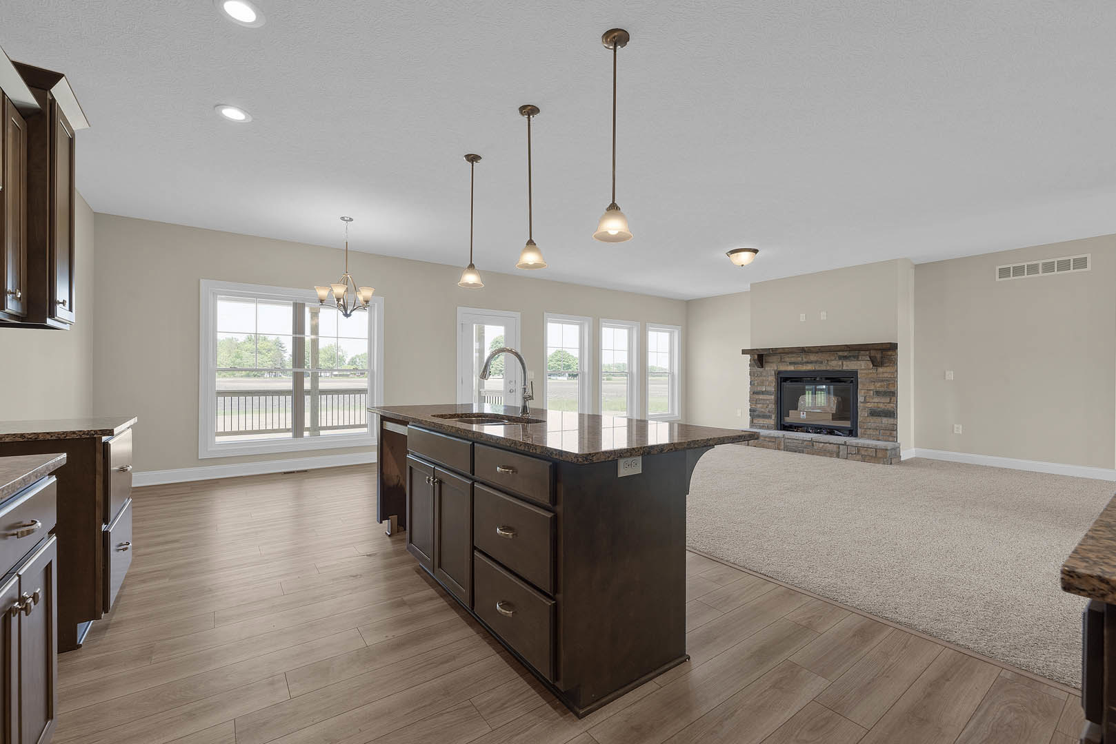 Open-concept kitchen and living room featuring a marble-topped island, white cabinetry, wood flooring, brick fireplace with firewood box, and large window overlooking the outdoors