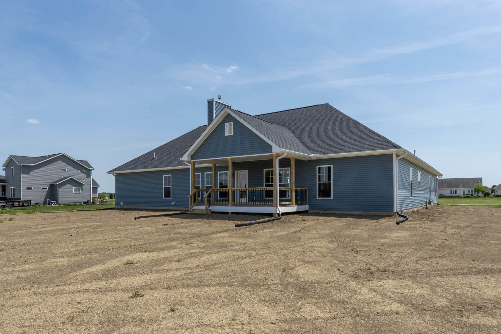 Single-story house with blue roof and white-framed windows, covered front porch, dirt field in foreground, Southfork Ranch visible in background under partly cloudy sky