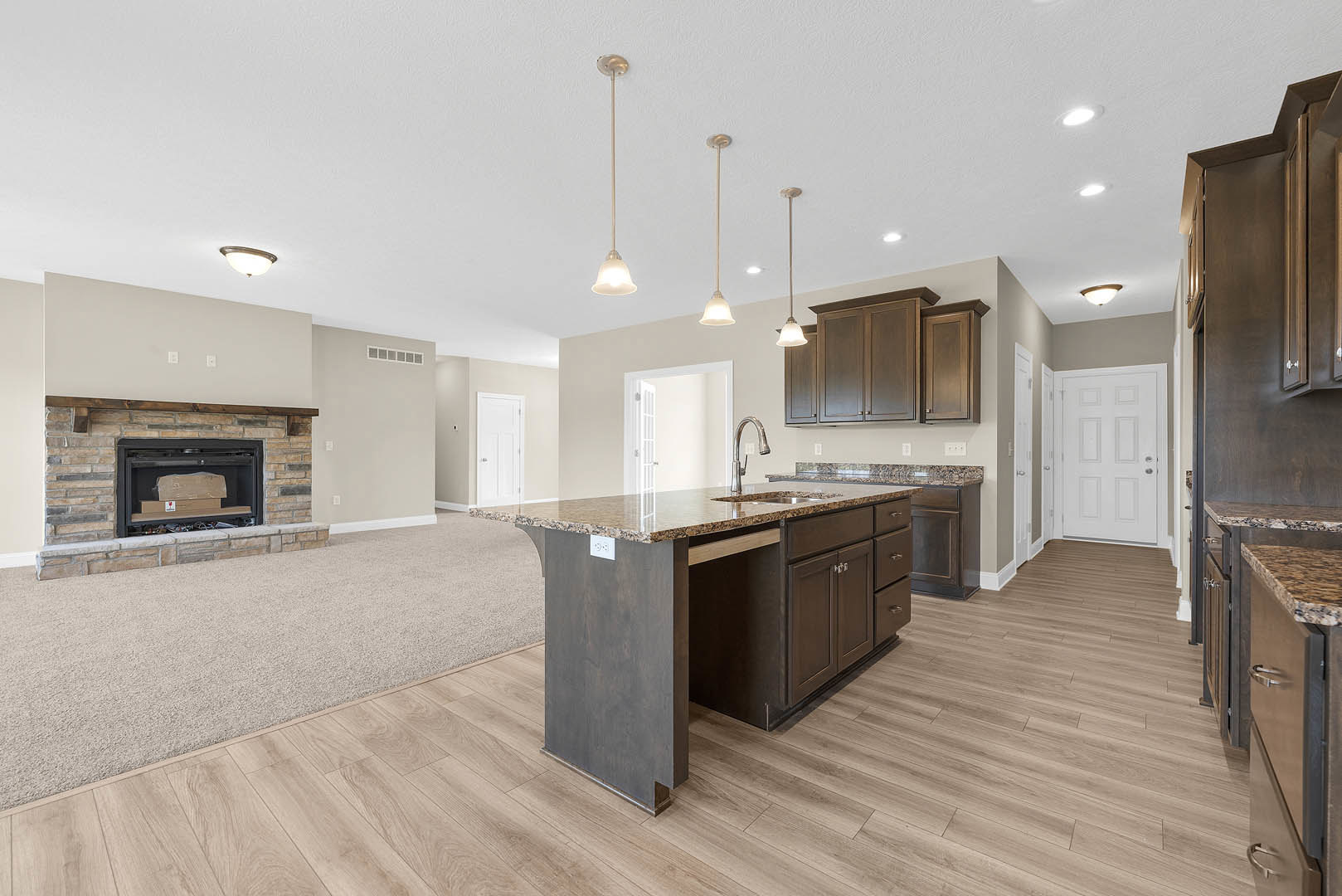 Open-concept kitchen and living room featuring a stone fireplace with a firewood box, white cabinetry, stainless steel handles, large kitchen island with built-in sink, wood