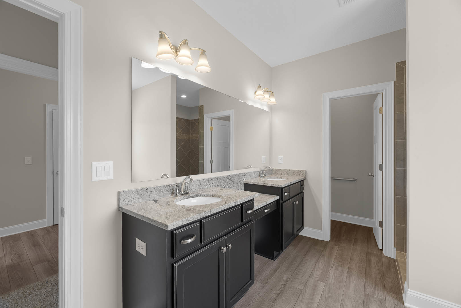 Bathroom with double sinks set in a stone countertop, large frameless mirror above, three-light fixture mounted on wall, white cabinetry, tile flooring, white door with gold hinge