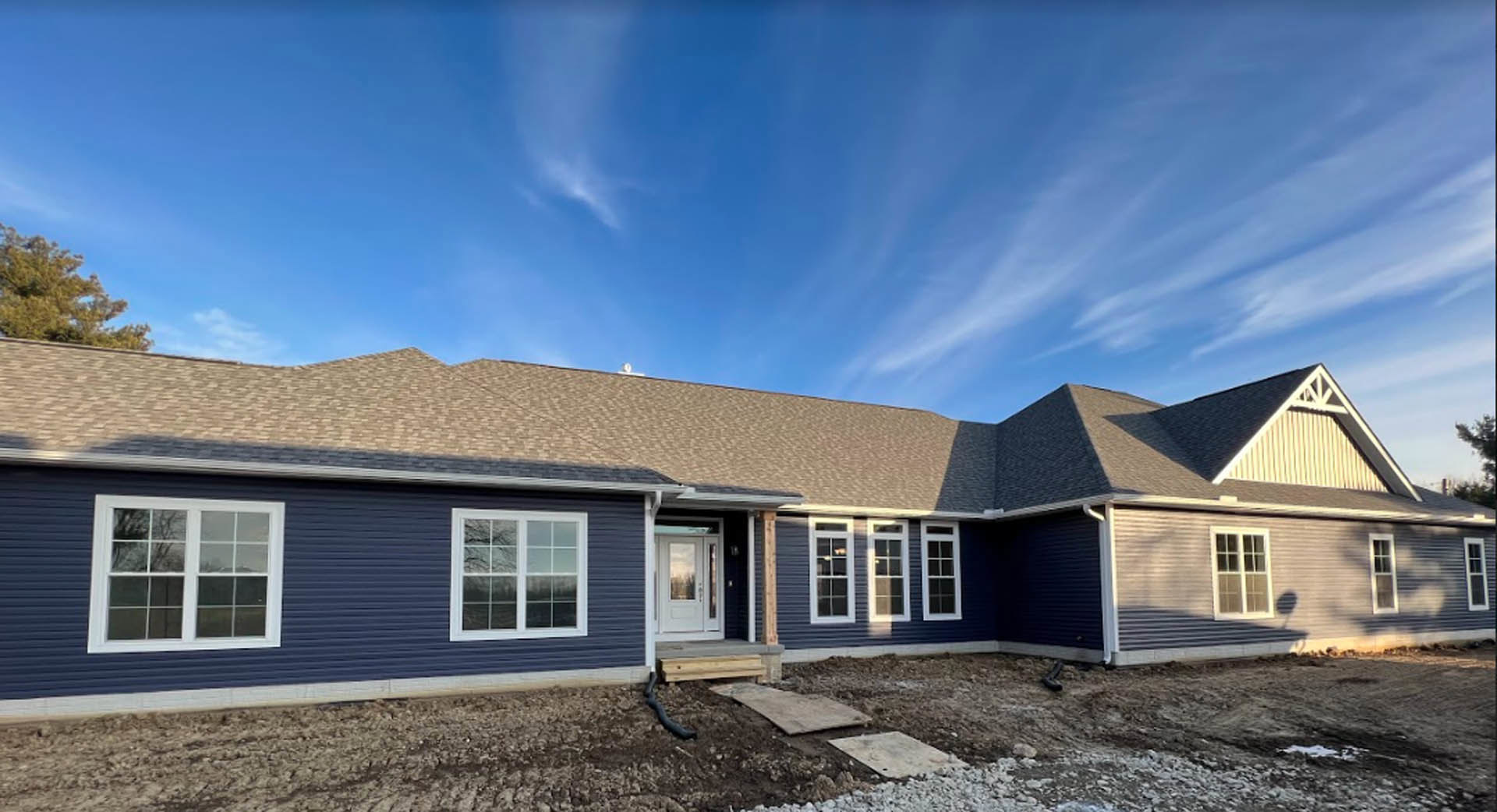 Framed house under construction with blue siding, white door and window, pitched roof, green tree, and clear blue sky