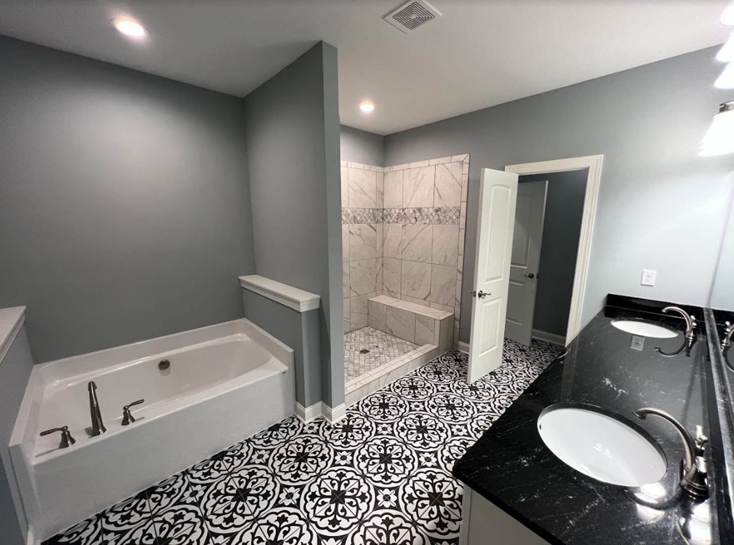 White freestanding bathtub with chrome faucets, black and white patterned tile floor, white sink set in black countertop, glass shower enclosure with built-in bench, open door