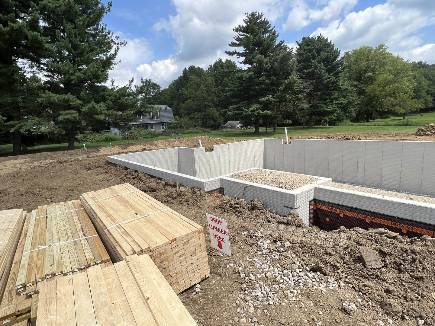 Concrete foundation surrounded by dirt and gravel, stacks of wood planks, pile of rocks, house under construction, red sign, trees, and blue sky in background